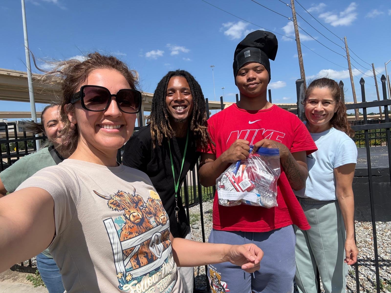 Group of five diverse people standing outdoors on a sunny day, smiling, with a black fence and utility poles in the background, taking a selfie.