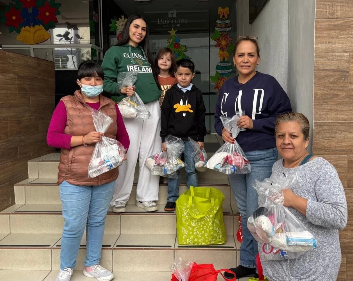 Group of six people standing on steps outside a building, holding bags of donated food and supplies, with Christmas decorations in the background.