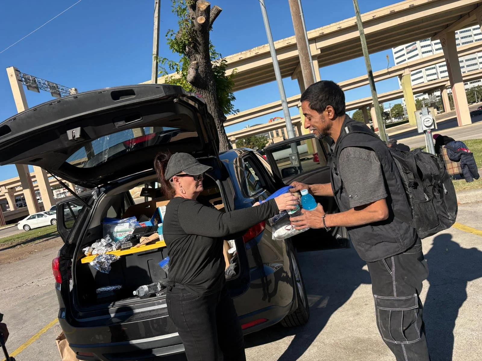 A woman handing a water bottle to a man near a black SUV with the trunk open, under a clear blue sky, with a cityscape and highway overpasses in the background.