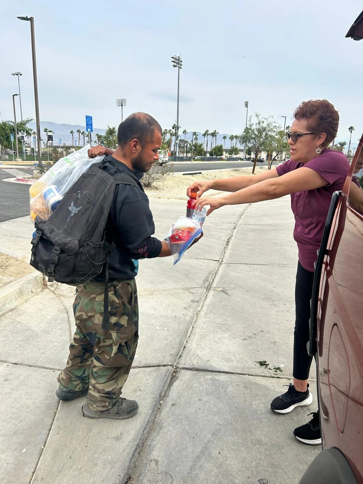 A woman in a purple shirt hands a bottle of water and a small item to a man with a backpack, standing on a sidewalk near a parked vehicle, under an overcast sky.
