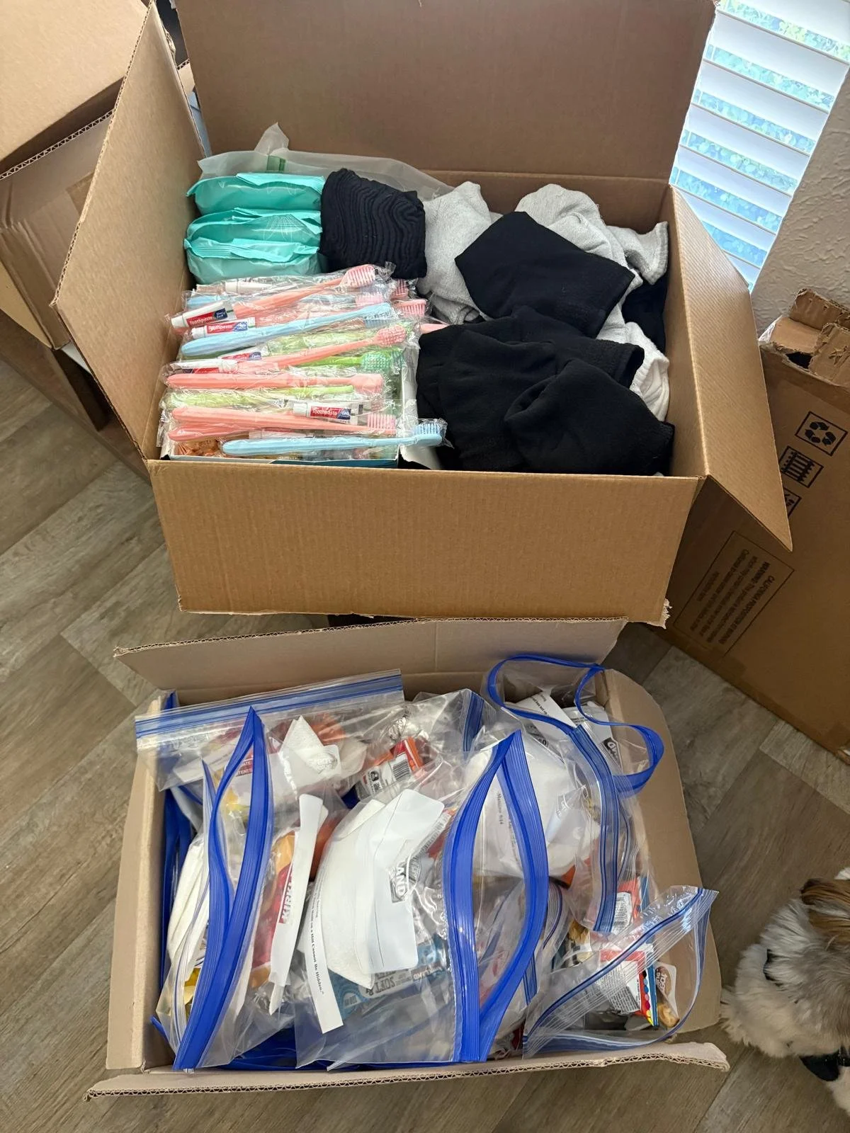 Two open cardboard boxes containing donated clothing and toiletries. The top box has folded clothes and toothbrushes in plastic packs. The bottom box has toiletry items in clear plastic bags.