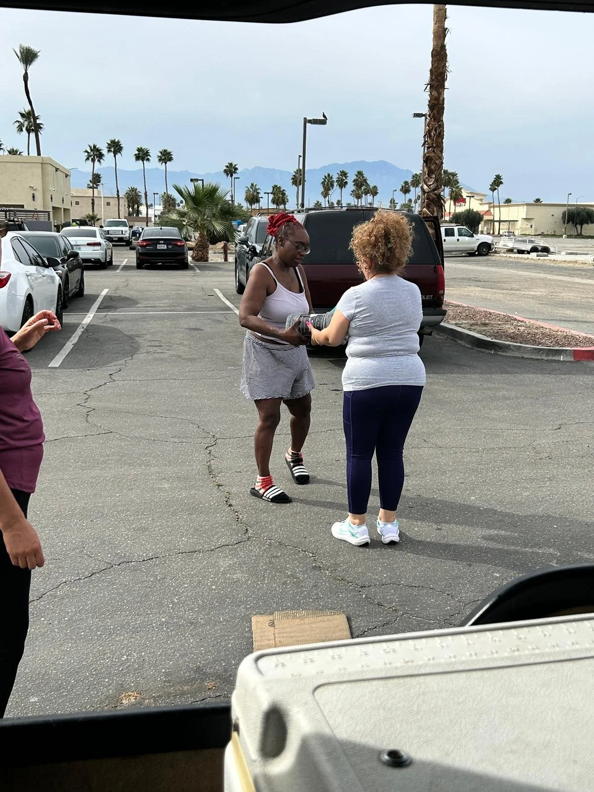Two women are exchanging a small gift or item in a parking lot with palm trees and mountains in the background. One woman has red hair and is wearing striped shorts and slides, while the other has curly blonde hair and is wearing a gray shirt and dar
