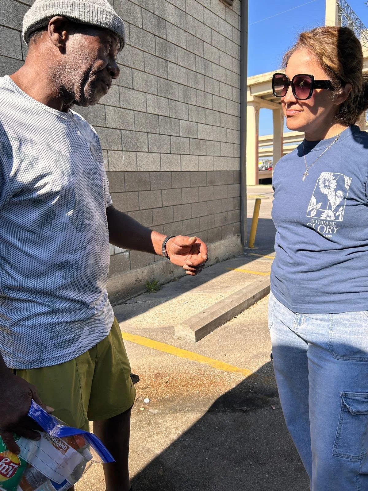 Two women are standing outside near a building, engaged in conversation. The woman on the left is wearing a gray beanie, a gray and white mesh shirt, olive shorts, and holding a snack bag. The woman on the right is wearing large sunglasses, a blue T-