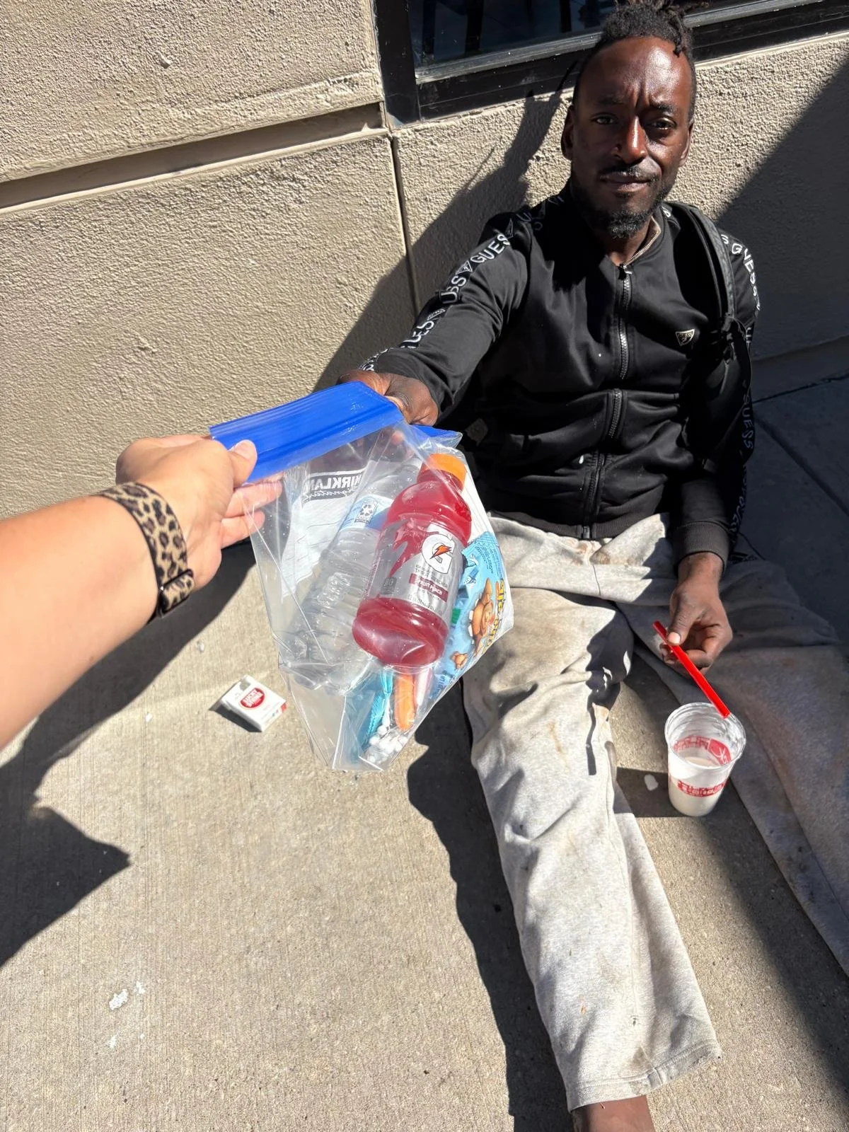 A man sitting on the sidewalk against a wall, holding a red Drink Gatorade bottle, while someone offers him a plastic bag containing water, a Gatorade drink, and a snack. The man is dressed in casual clothing and has a drink in a cup with a red straw.