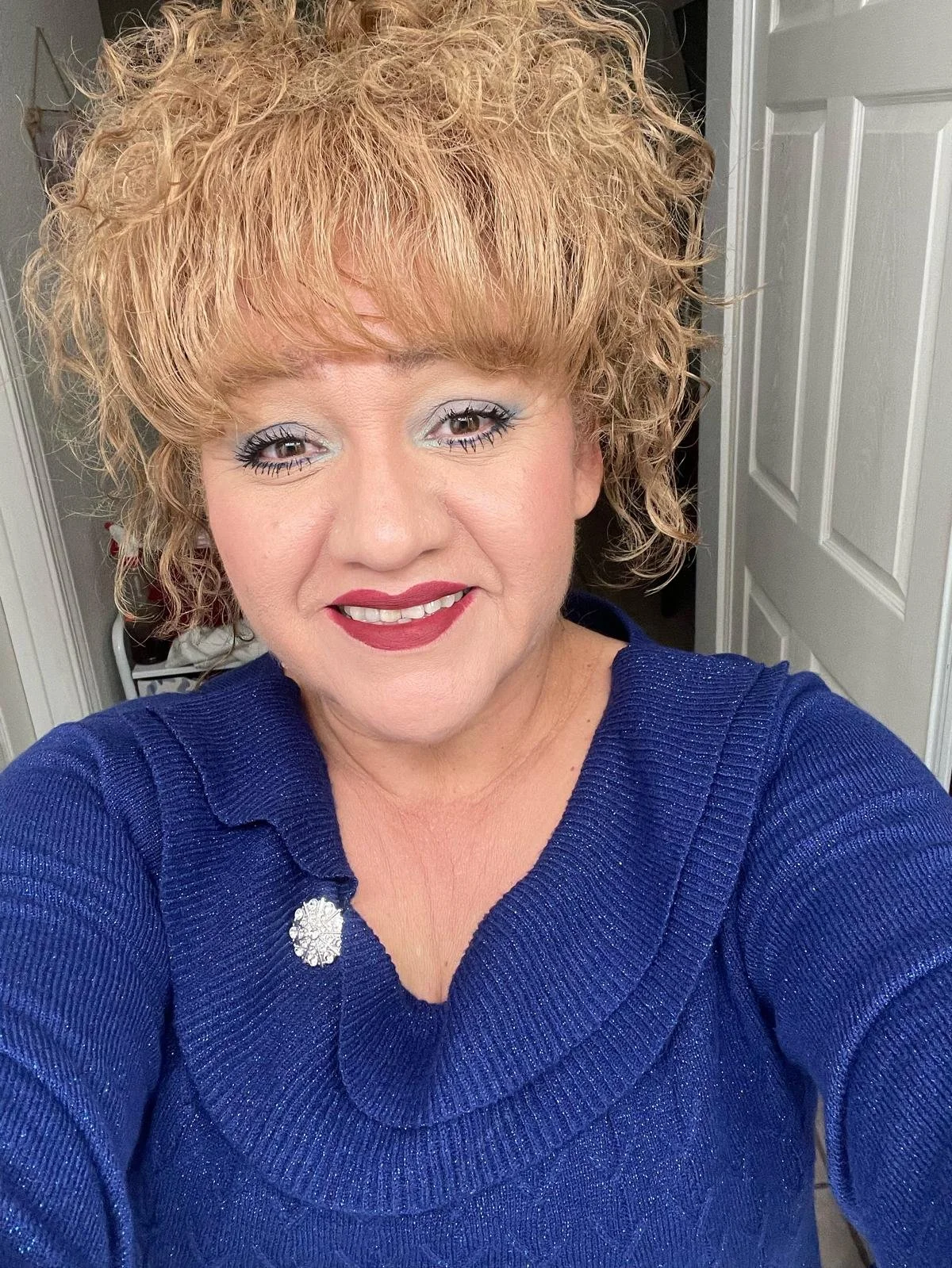 A woman with curly blonde hair taking a selfie indoors, wearing a navy blue sweater with a decorative clasp.