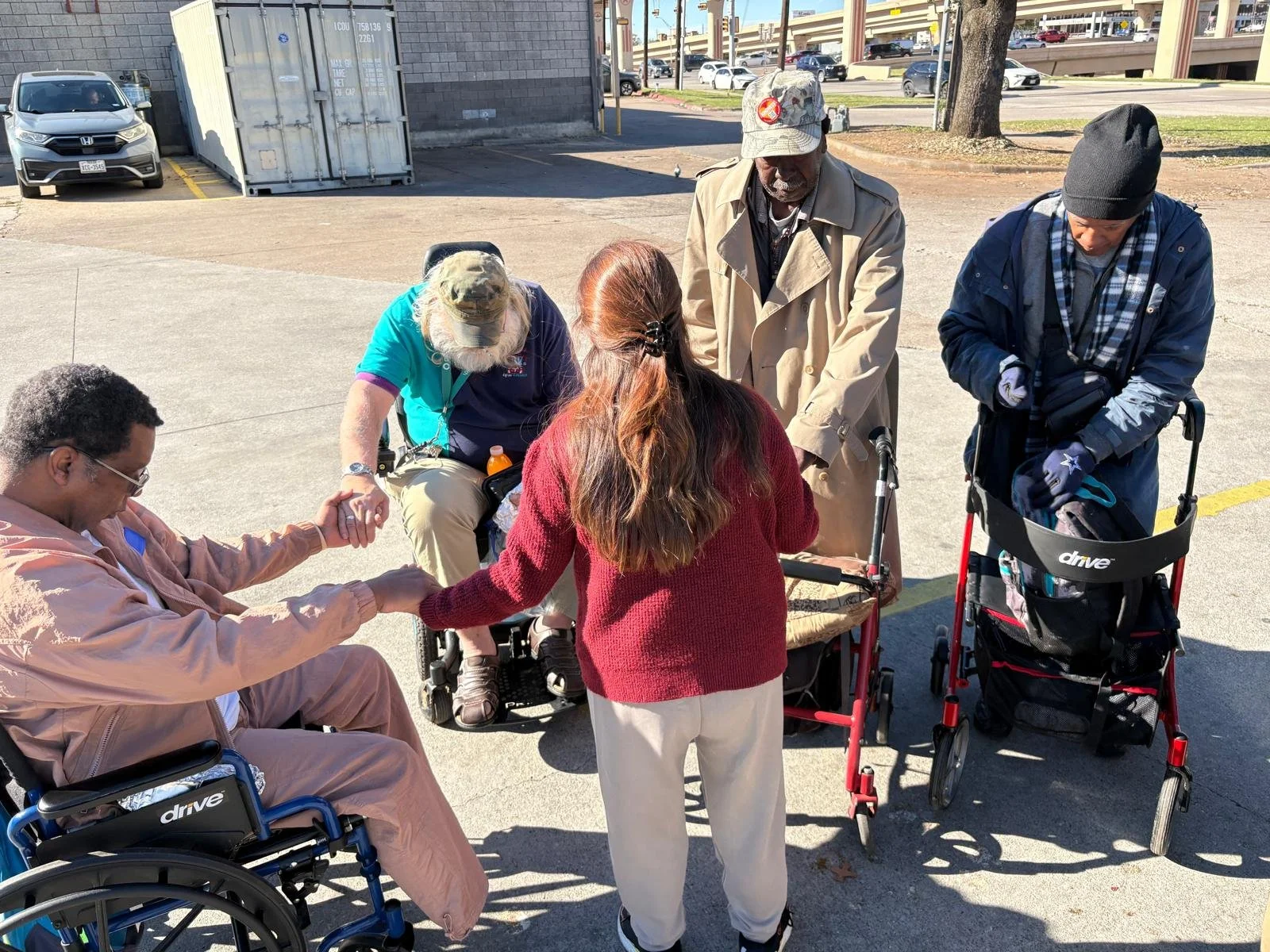 A group of five people, some in wheelchairs and some standing, holding hands in a circle outside on a sunny day. There are vehicles and a building in the background.
