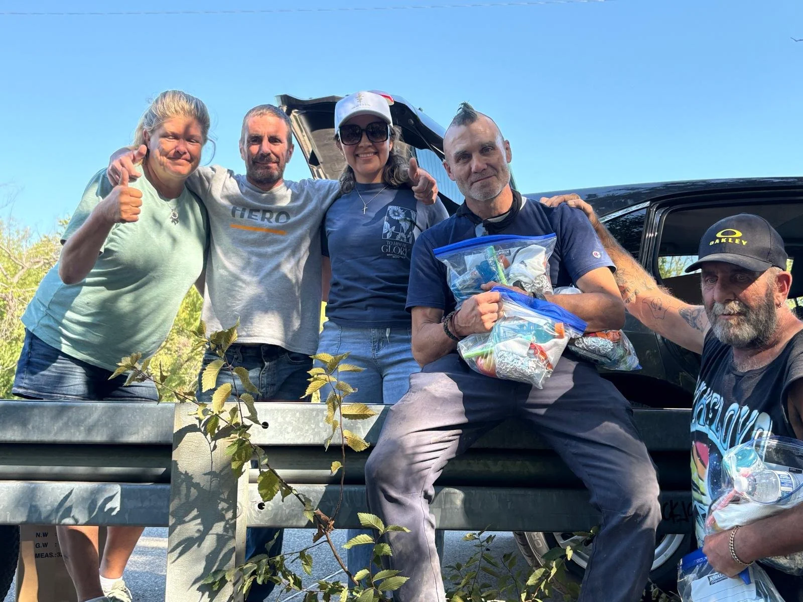 Group of five people outdoors near a black vehicle, with one holding plastic bags with supplies, smiling and posing for a photo on a sunny day.
