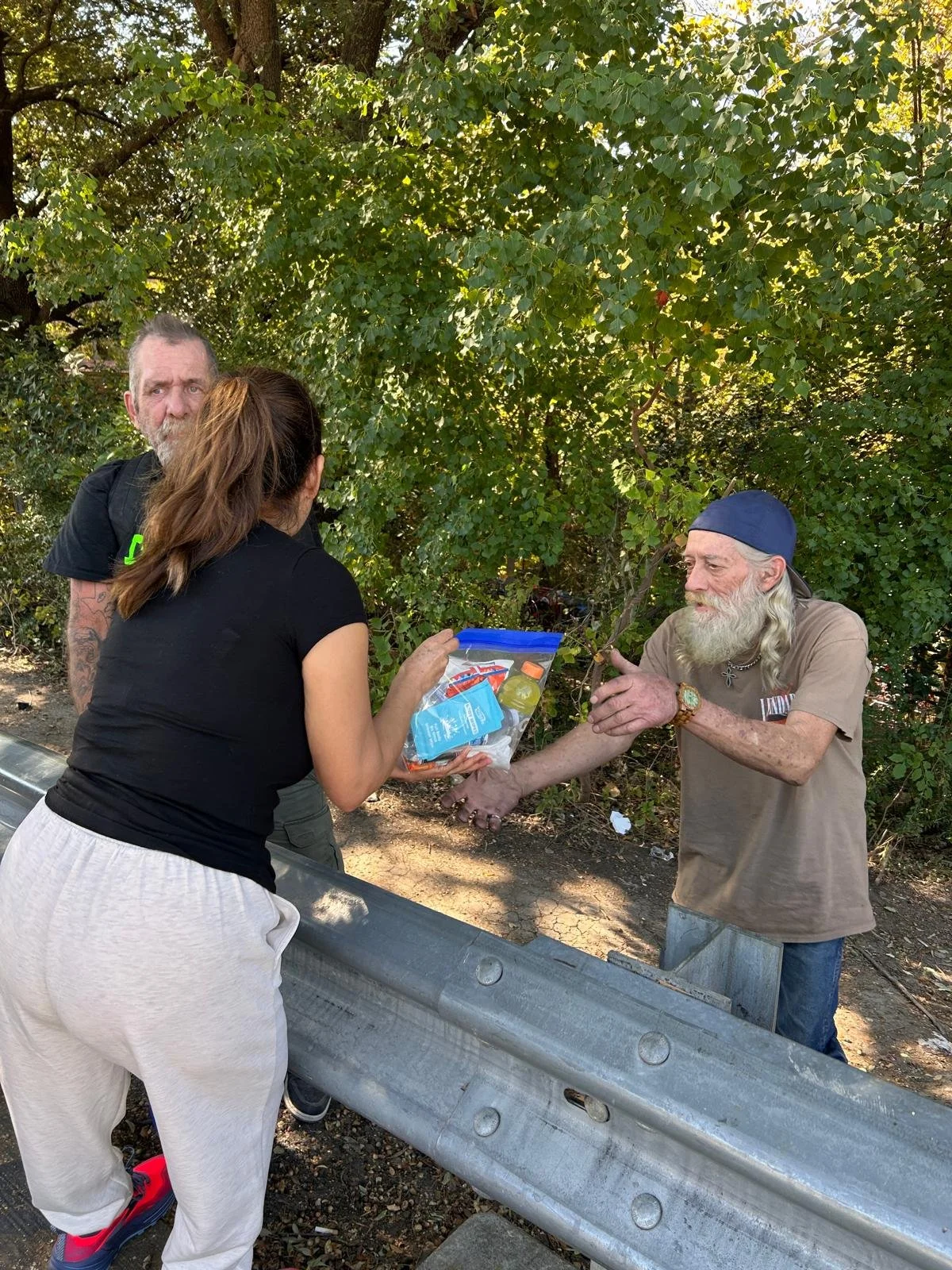 A woman is giving a bag of groceries to an elderly man with a long white beard, wearing a blue bandana. Two other people, one man and one woman, stand nearby on a wooded roadside.