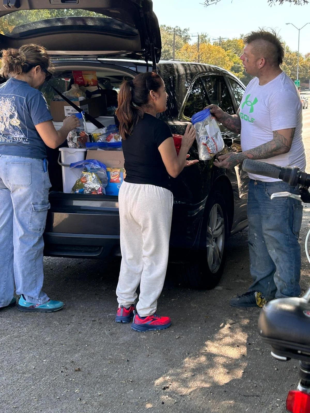 Three people loading groceries into the trunk of a black SUV on a sunny day in a parking lot, with trees and power lines in the background.