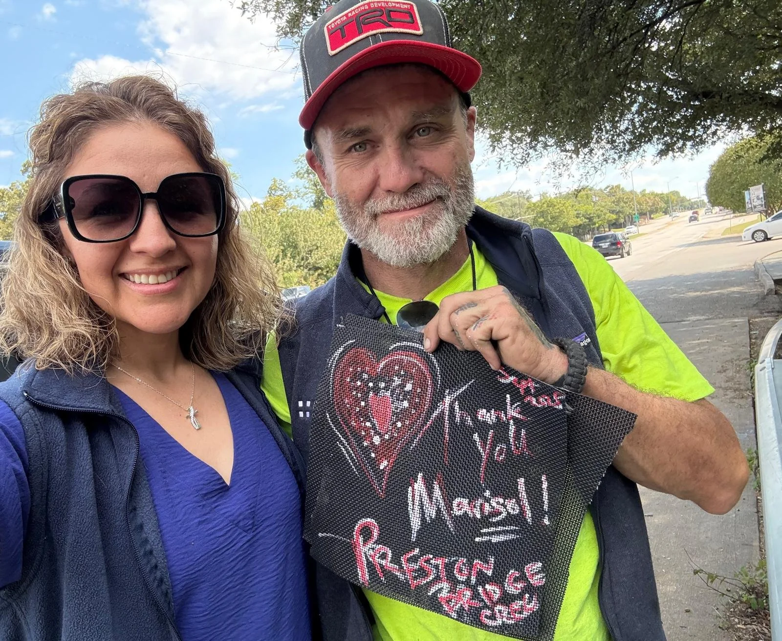 A woman with sunglasses and a man with a beard, wearing a red and black cap, holding a sign with a heart and handwritten message, standing outdoors on a sunny day with trees and parked cars in the background.