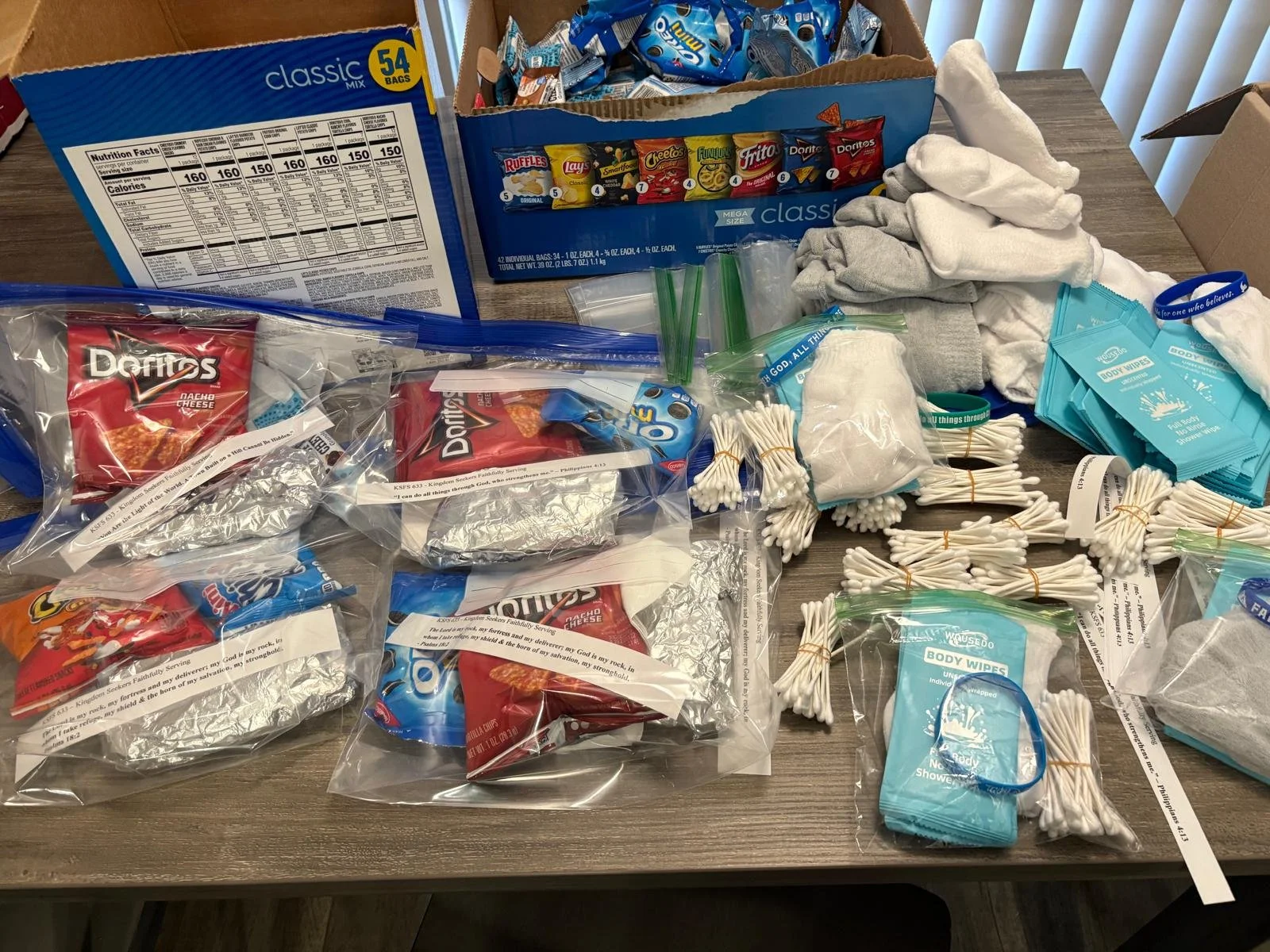 A table filled with sanitation supplies including packaged snack foods, cotton balls, bundles of cotton swabs, small bottles of hand sanitizer, folded socks, face masks, and unopened packs of body wipes.
