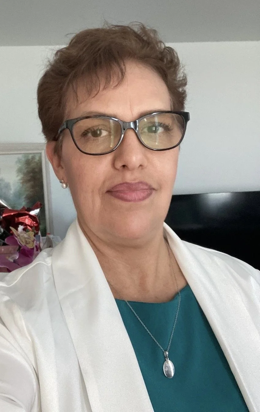 A middle-aged woman with short brown hair, wearing glasses, a teal top, a white blazer, and a pendant necklace.