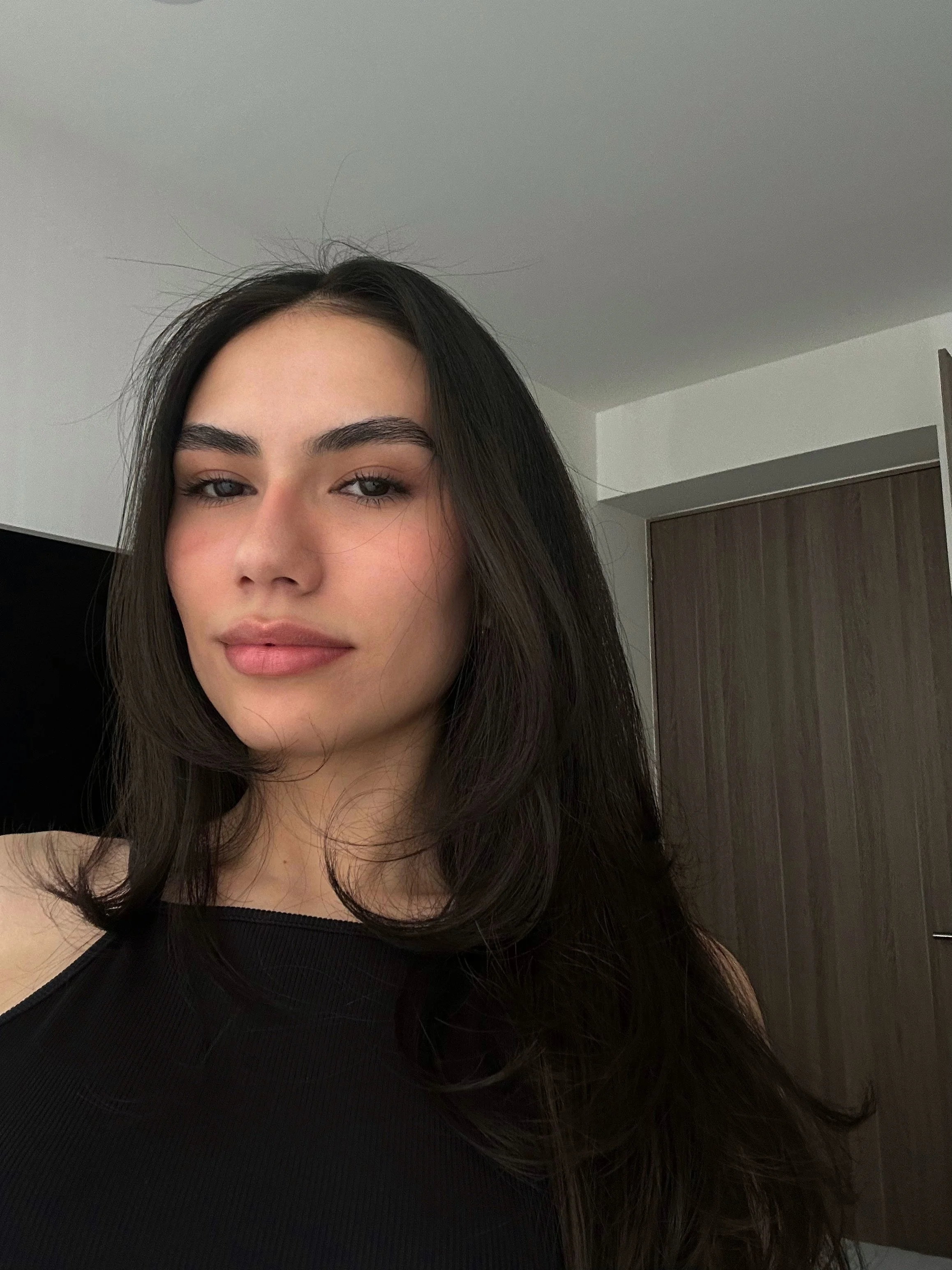 A young woman with dark hair and natural makeup taking a selfie indoors.