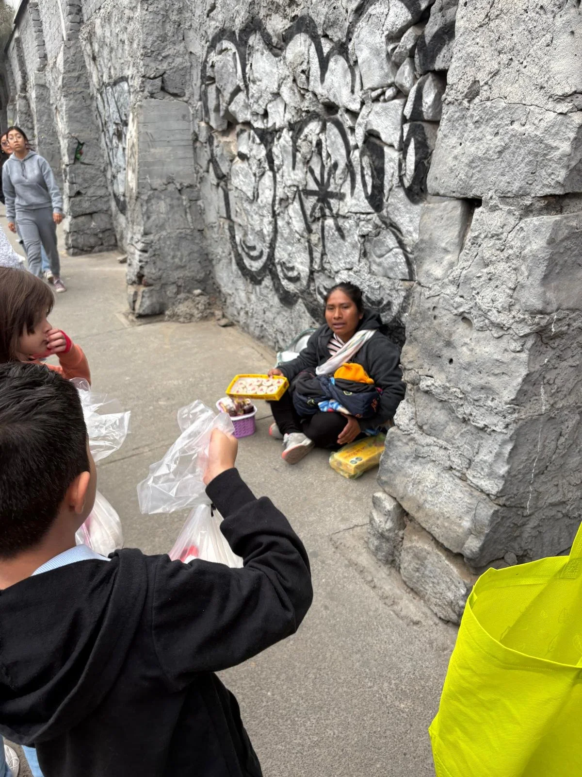 A woman sitting on the ground next to a large stone wall with graffiti, selling snacks to a group of children and adults nearby.