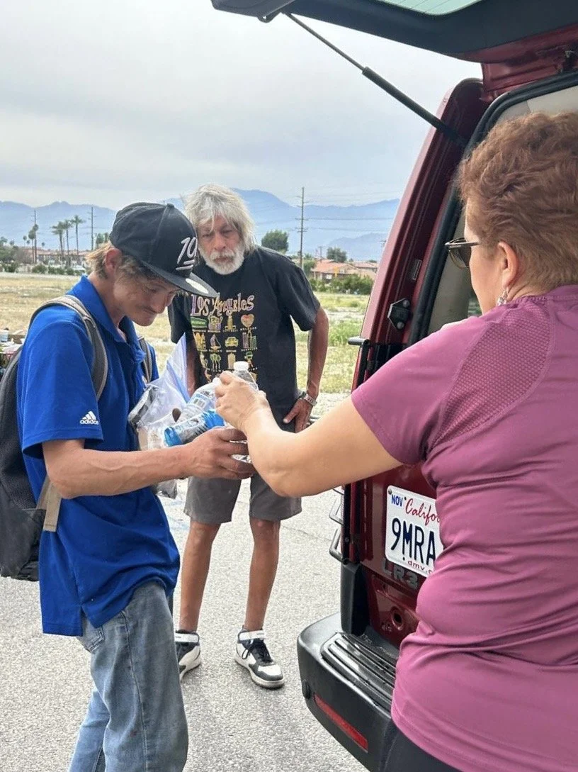 A person receiving water from a woman at the back of a red vehicle with a California license plate, while another man watches.