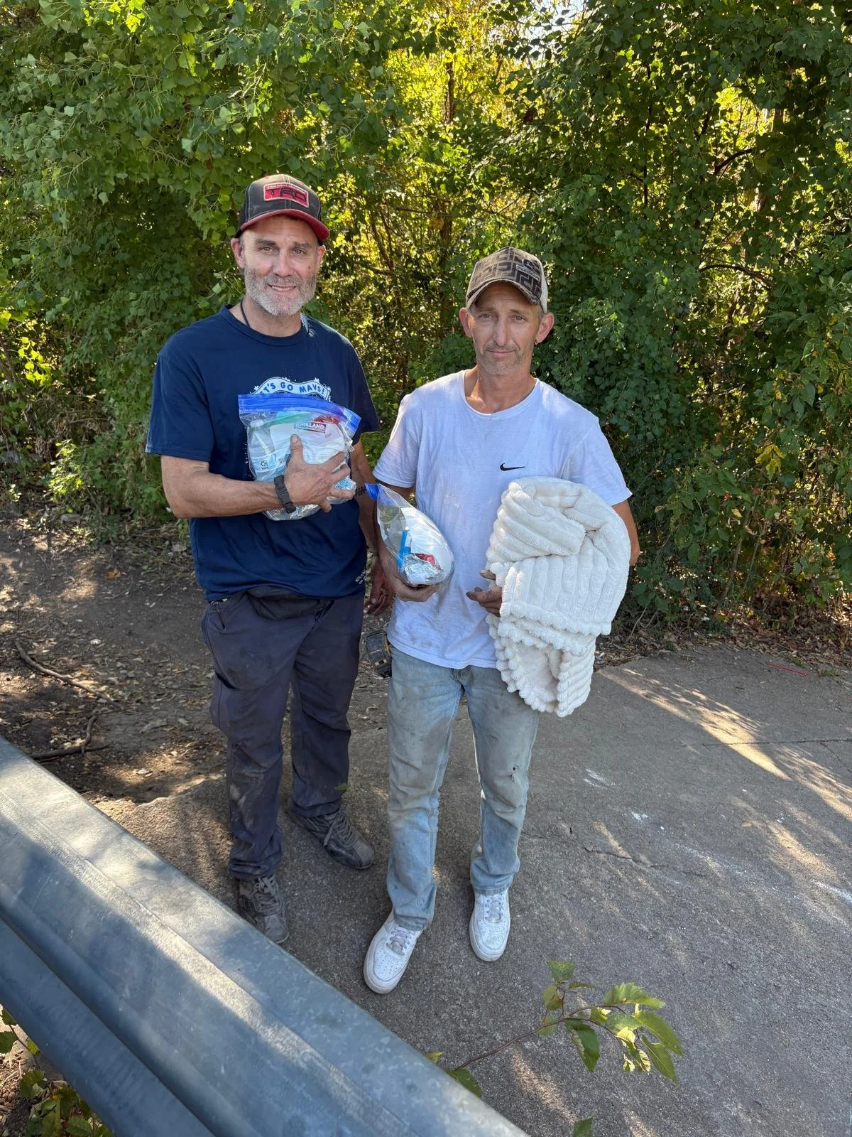 Two men standing outdoors on a paved path, holding supplies and a blanket, with green trees and bushes in the background.
