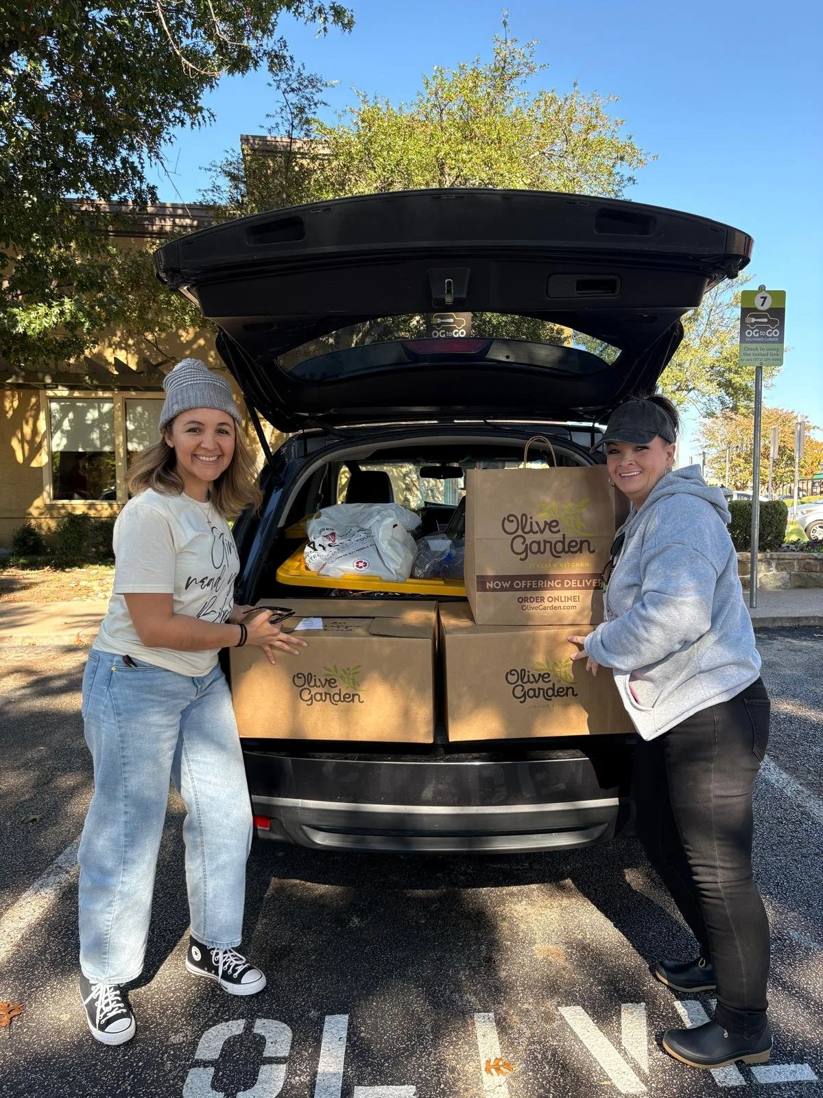 Two women load grocery bags into the trunk of a car in a parking lot on a sunny day.