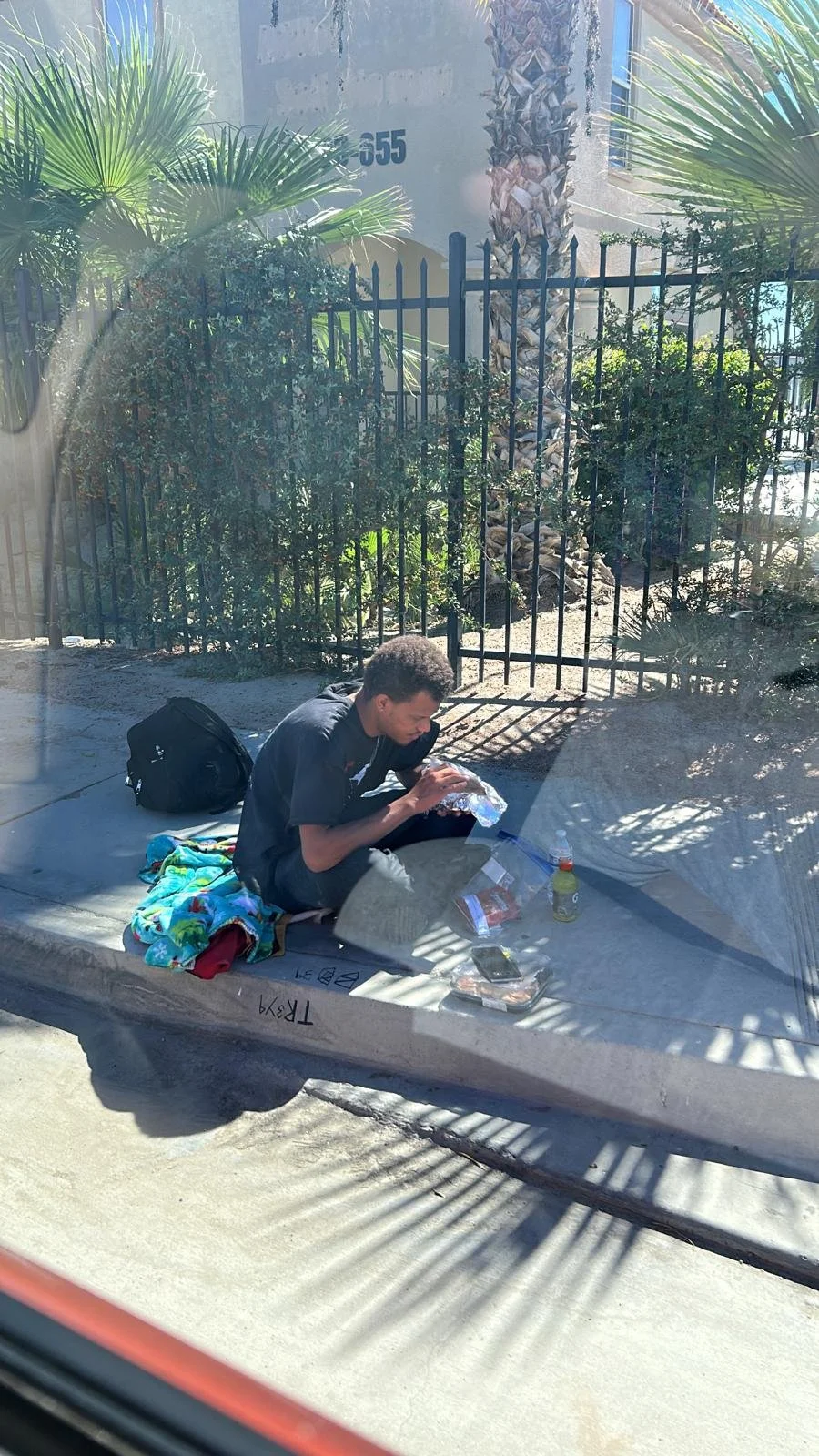 A young man sits on a sidewalk at a bus stop, eating food from a foil container with bottled drinks nearby. He has a backpack and a colorful blanket or towel next to him, with a fence, trees, and a building in the background.