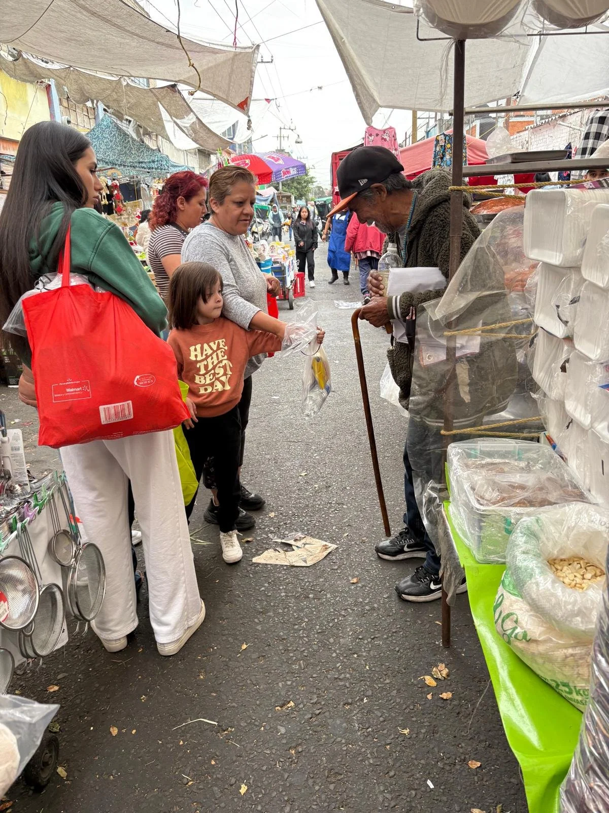 A man with a walking cane is selling items at a street market, while four women and a young girl are shopping and observing. The market is covered with tents and umbrellas on a busy street with other vendors and shoppers in the background.
