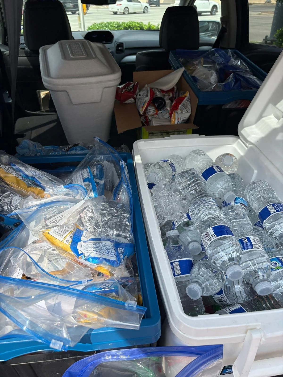 The trunk of a car filled with donated water bottles, plastic bags, and boxes of snacks and supplies.