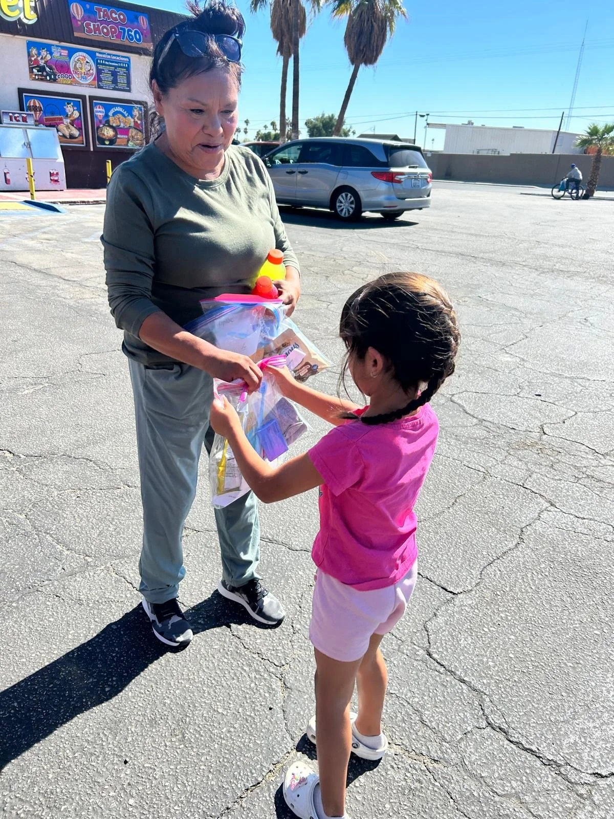 A woman and a girl standing in a parking lot, exchanging a plastic bag with various items, with cars and a taco shop in the background.