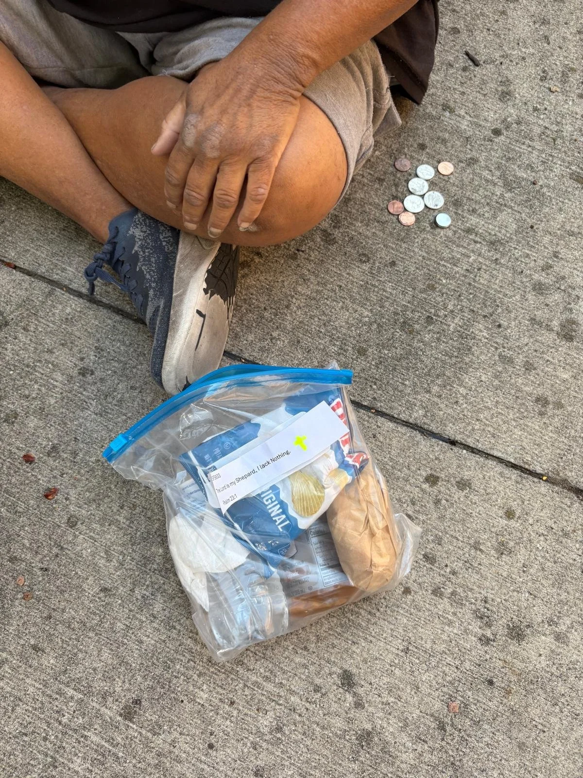 A person sitting on a sidewalk with a bag of food and some coins nearby.