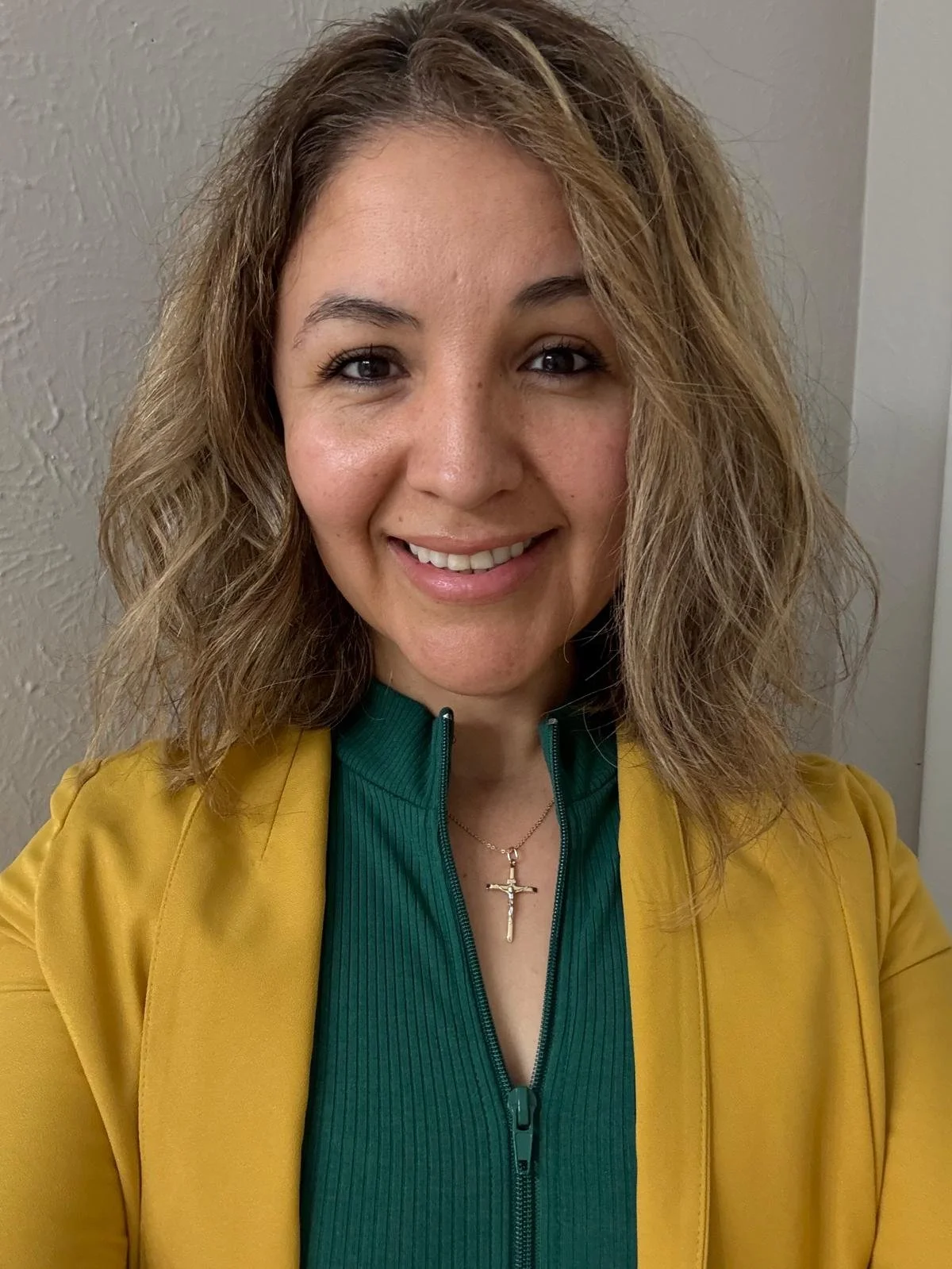 Close-up of a young woman with wavy, light brown hair, smiling, wearing a green zip-up top, a yellow blazer, and a gold cross necklace, standing indoors against a neutral-colored wall.
