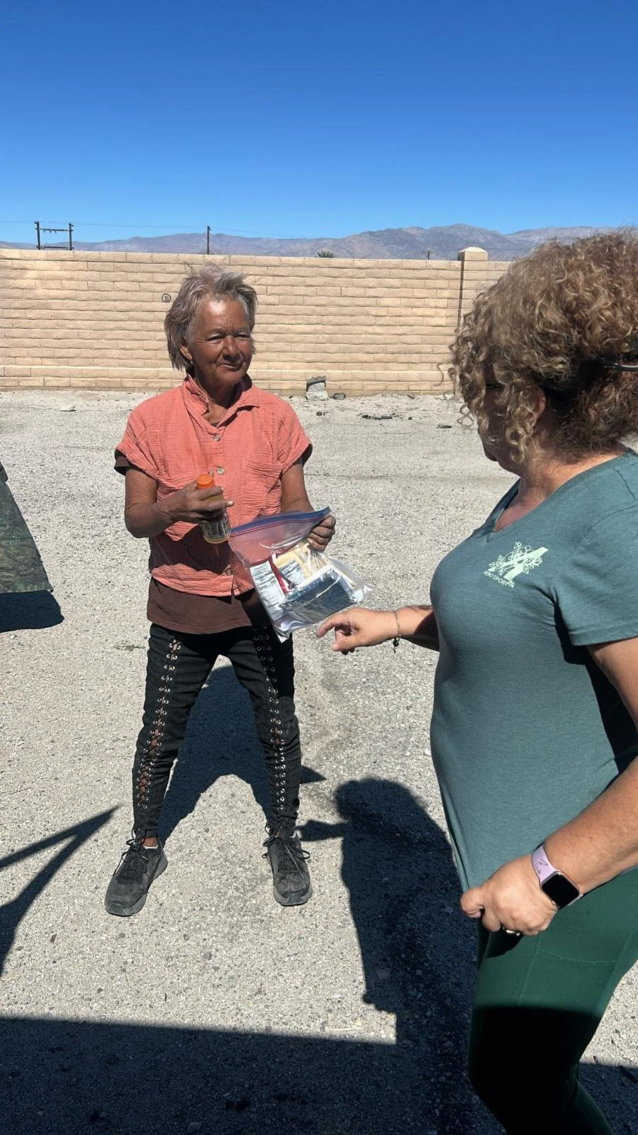 Two women are outdoors on a sunny day, one holding a plastic bag with various items and the other pointing at it. They are standing on a gravel surface with a beige brick wall and mountains in the background.