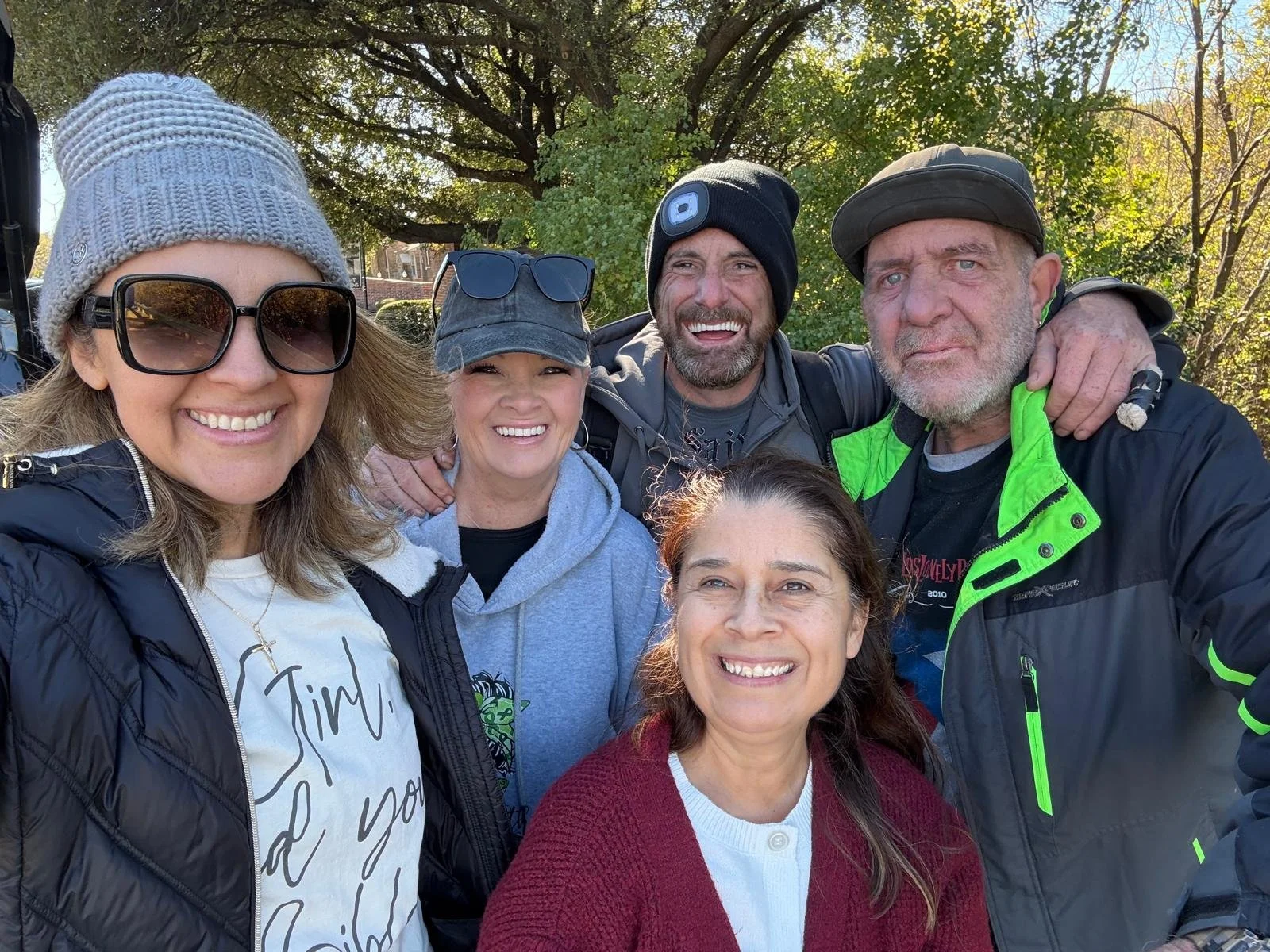Group of five people smiling outdoors, backdropped by trees, in a park-like setting with sunlight filtering through leaves.