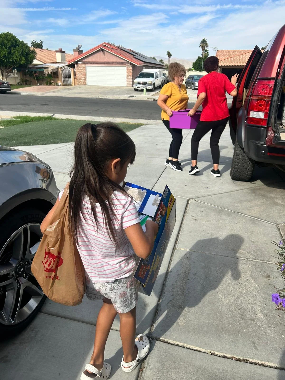 A young girl with long dark hair, wearing a striped pink and white shirt, floral shorts, and white Crocs, stands on a sidewalk holding a boxed toy with a brown paper bag over her shoulder. In the background, two women are loading or unloading items f