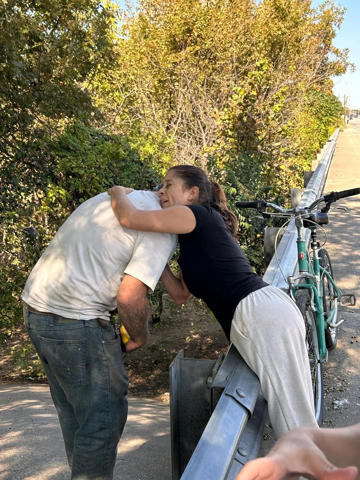 A woman hugging an older man on a hillside or rural area with trees, a bicycle and a guardrail in the background.