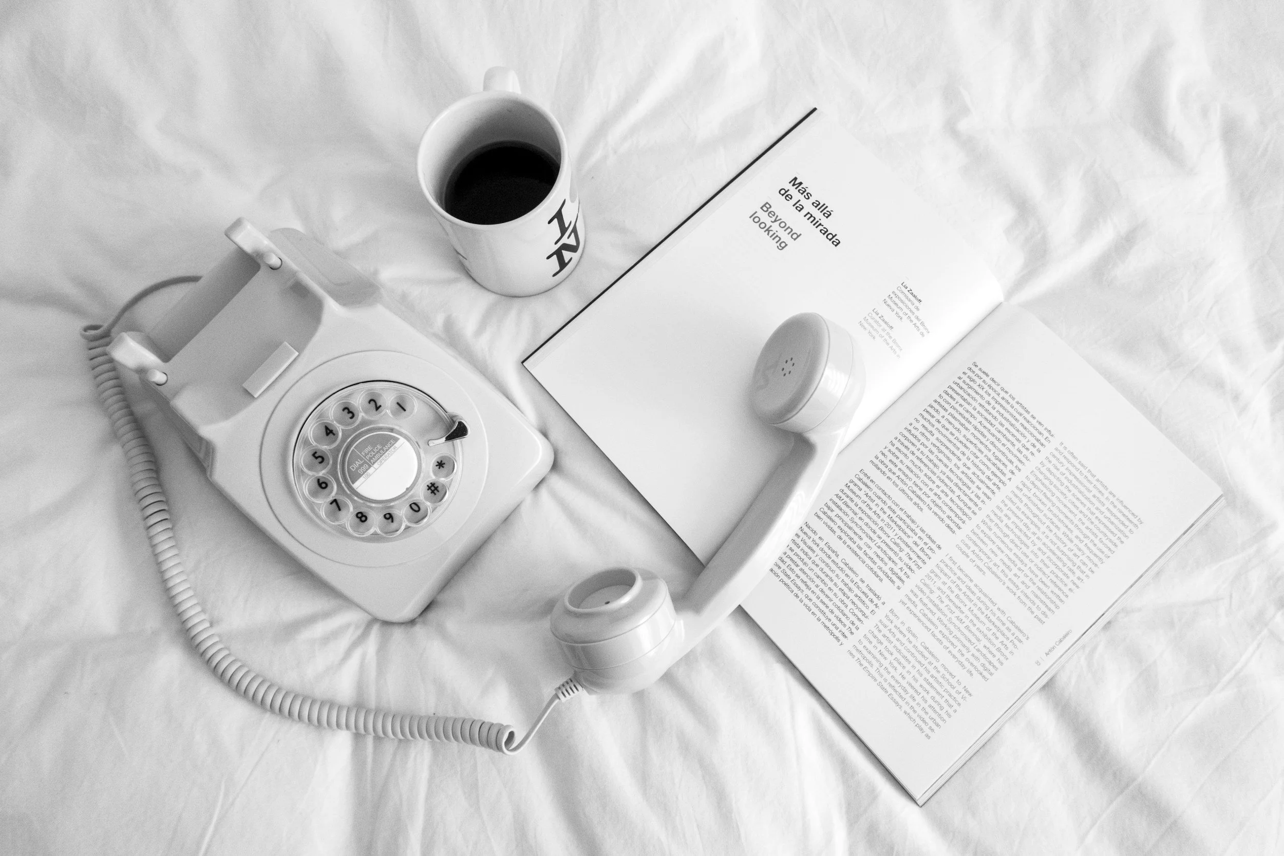 A black and white photograph of a vintage rotary telephone, an open magazine, a cup of coffee, and an air freshener on a bed with a white sheet.