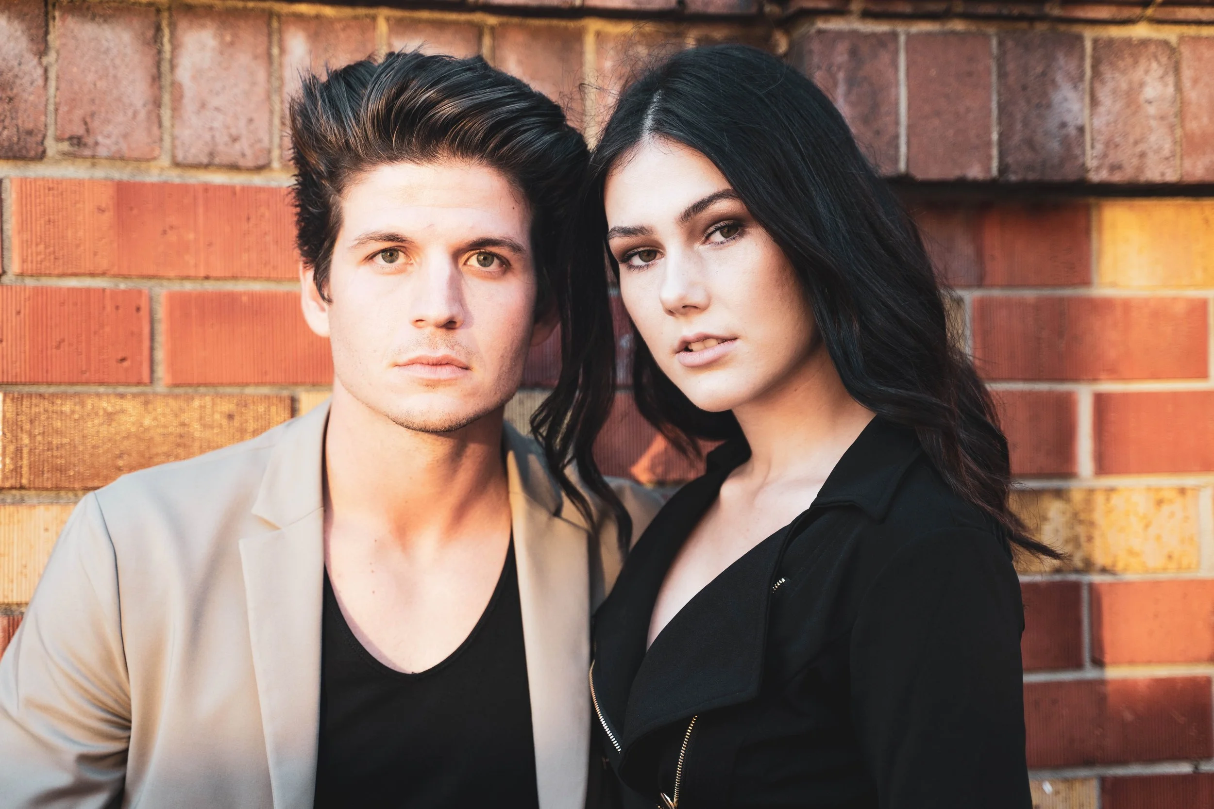 A young man and woman standing close together against a brick wall, looking at the camera.
