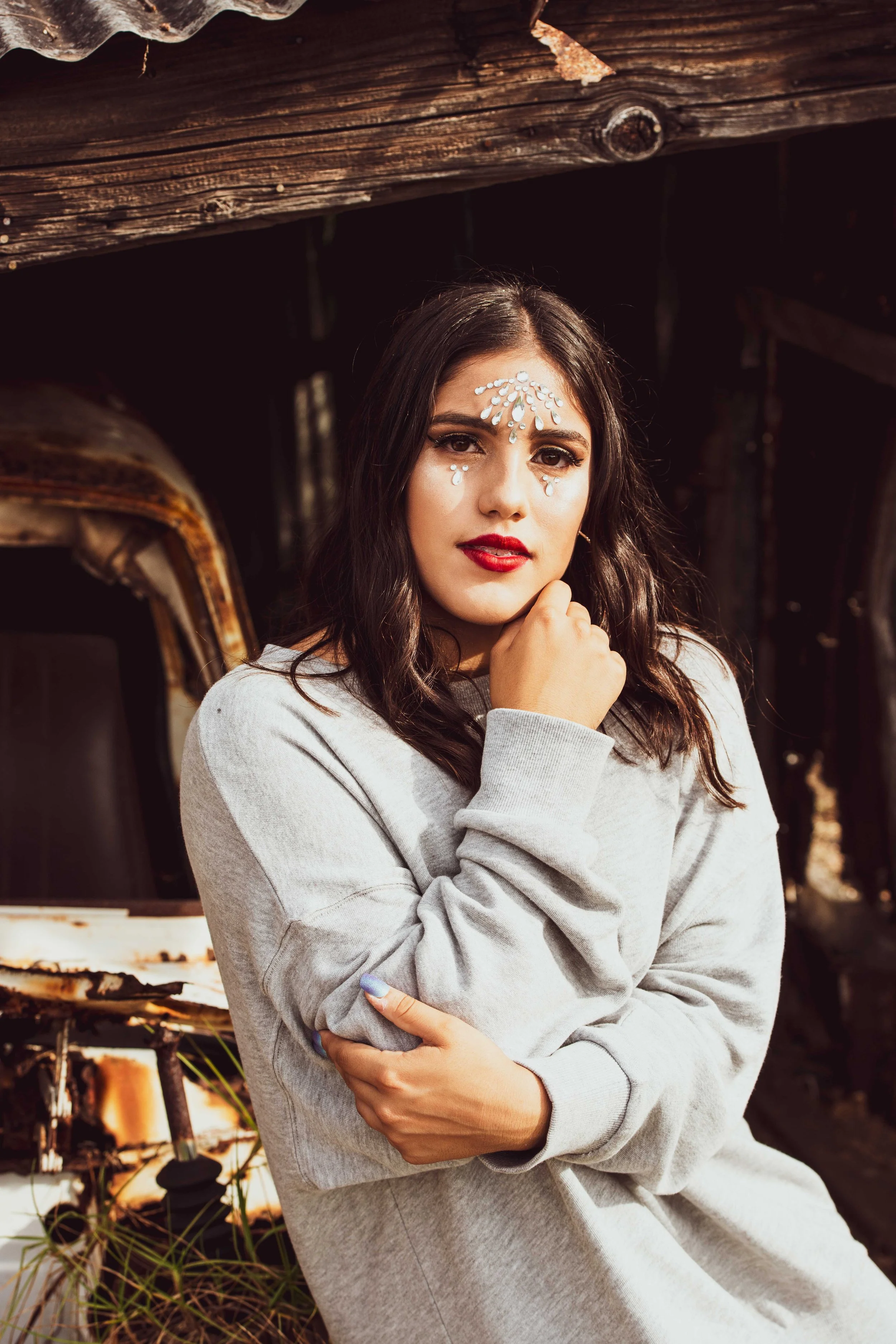 A young woman with medium-length dark brown hair and red lipstick poses outdoors, with sparkling rhinestones on her forehead and cheeks, wearing a light gray sweatshirt, in front of a rusty, abandoned vehicle and weathered wooden structure.