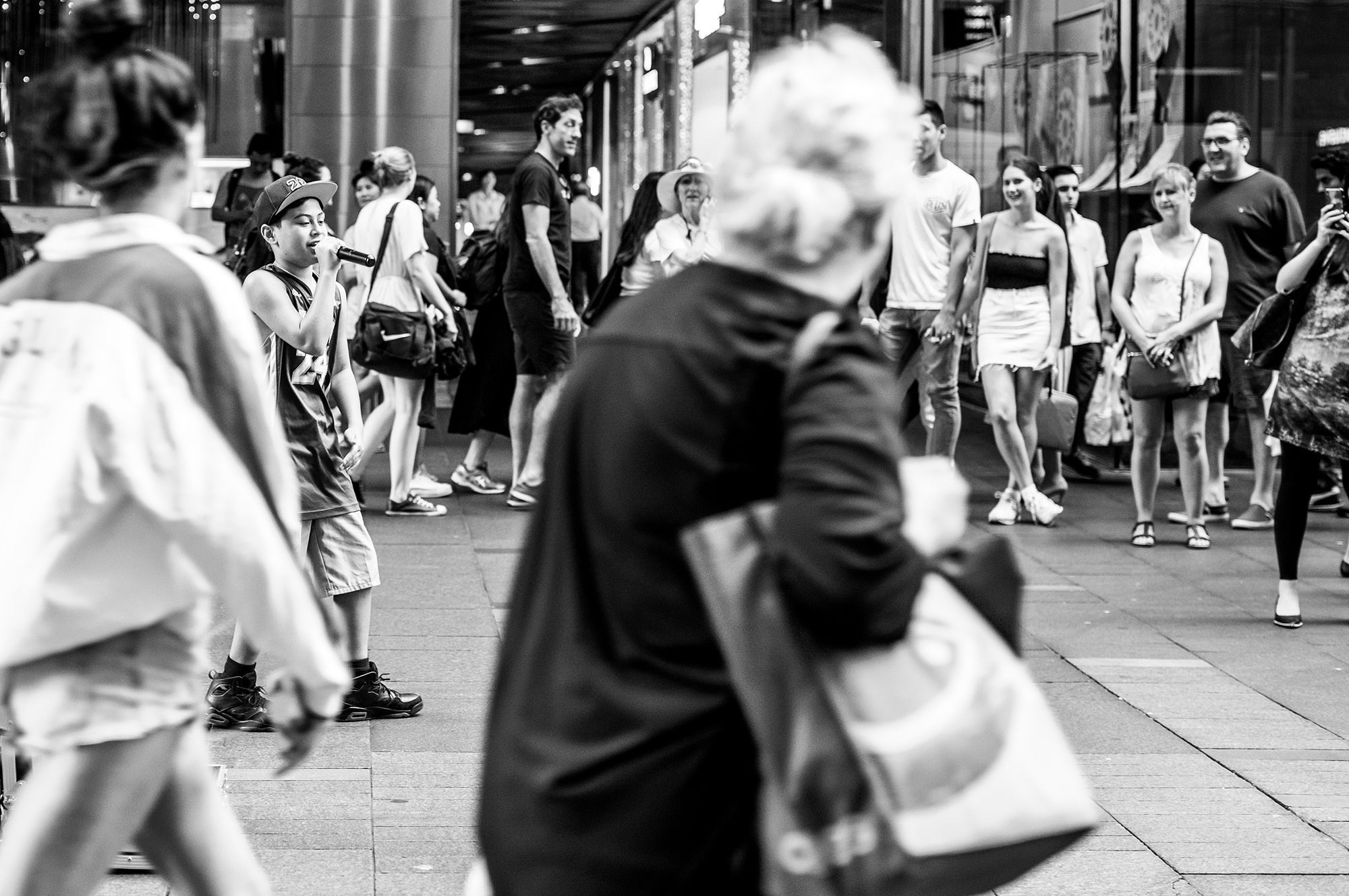 A girl singing into a microphone in a busy outdoor setting with people walking and standing around, some smiling and engaging.