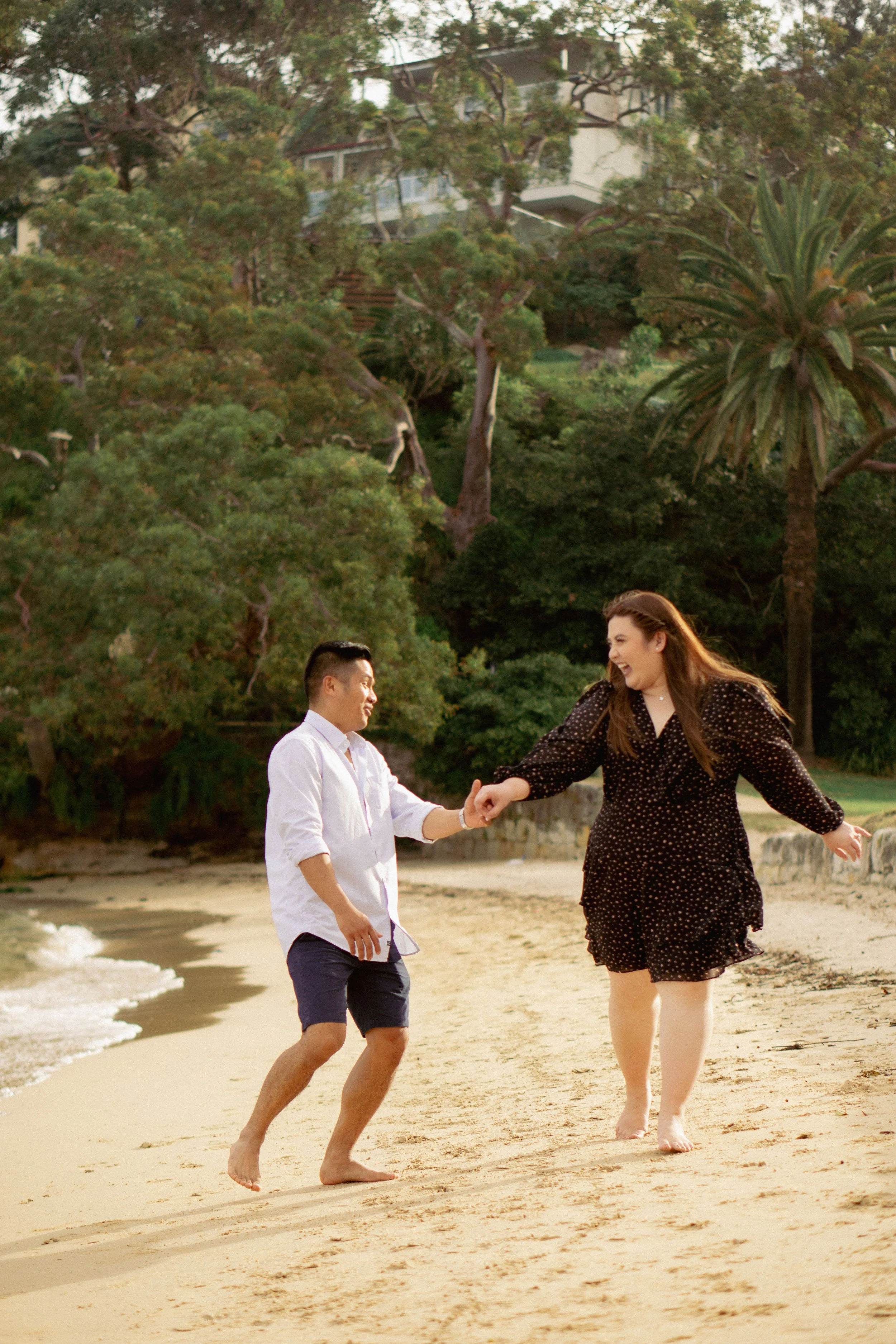 A couple laughing and holding hands while walking barefoot on a sandy beach near the shoreline, surrounded by greenery and trees.
