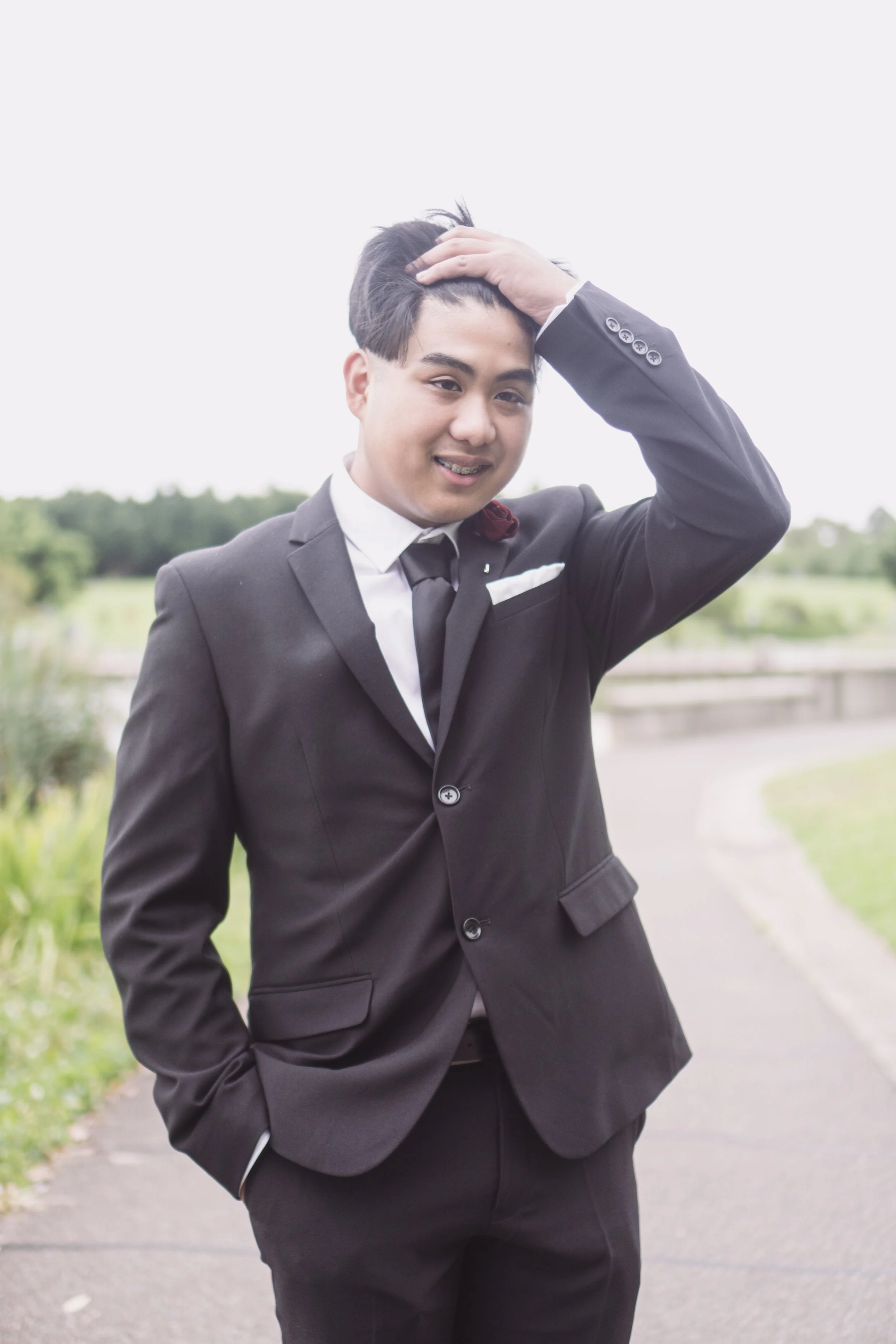 Young man in a black suit with a white shirt and black tie, standing outdoors on a pathway with greenery in the background, posing with one hand on his hair and smiling.