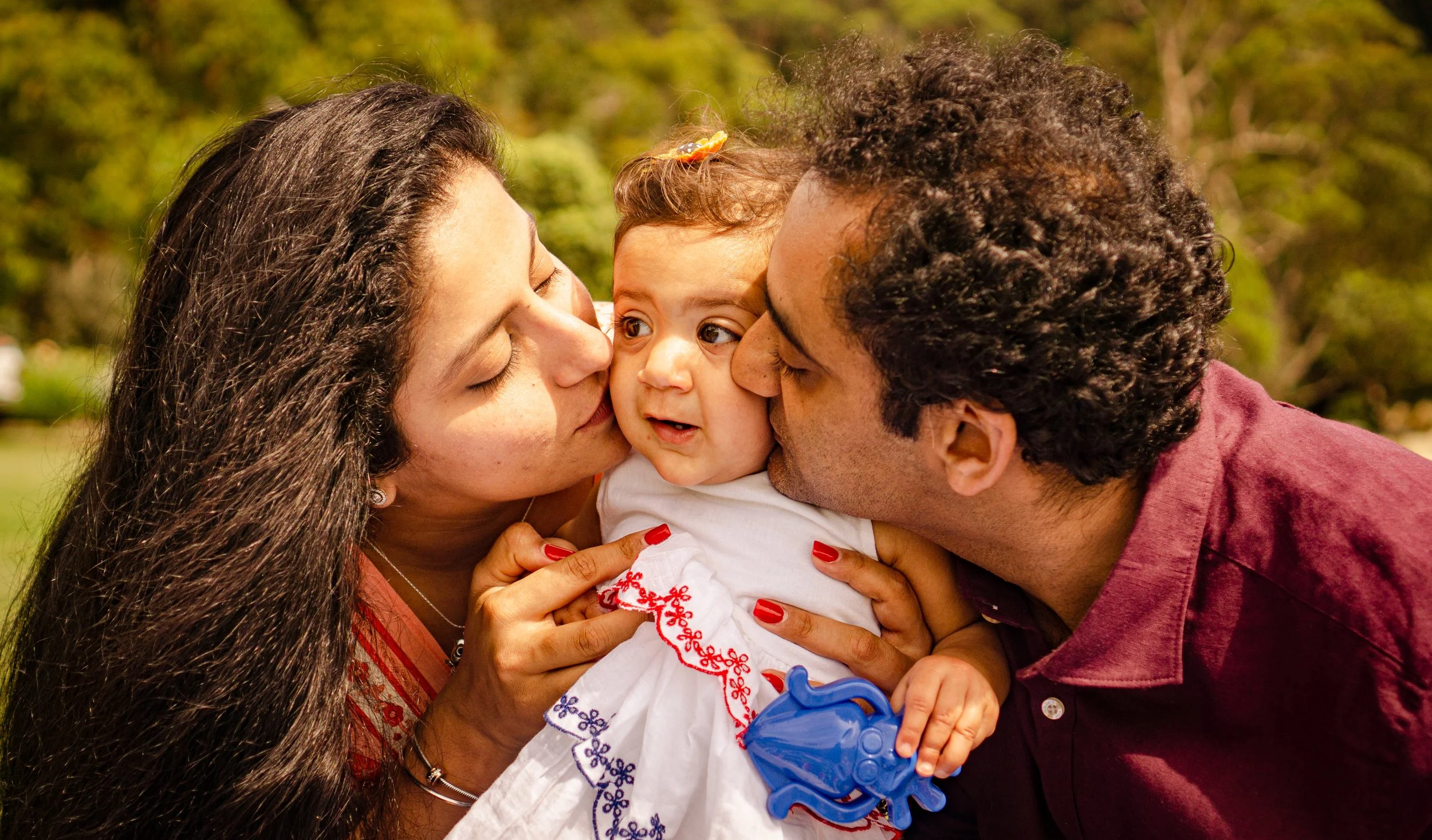 A family of three with light and dark skin tones, a woman, a man, and a baby, outdoors in a park, sharing a moment of affection. The woman and man are kissing the baby on the cheeks, and the baby is holding a blue toy.
