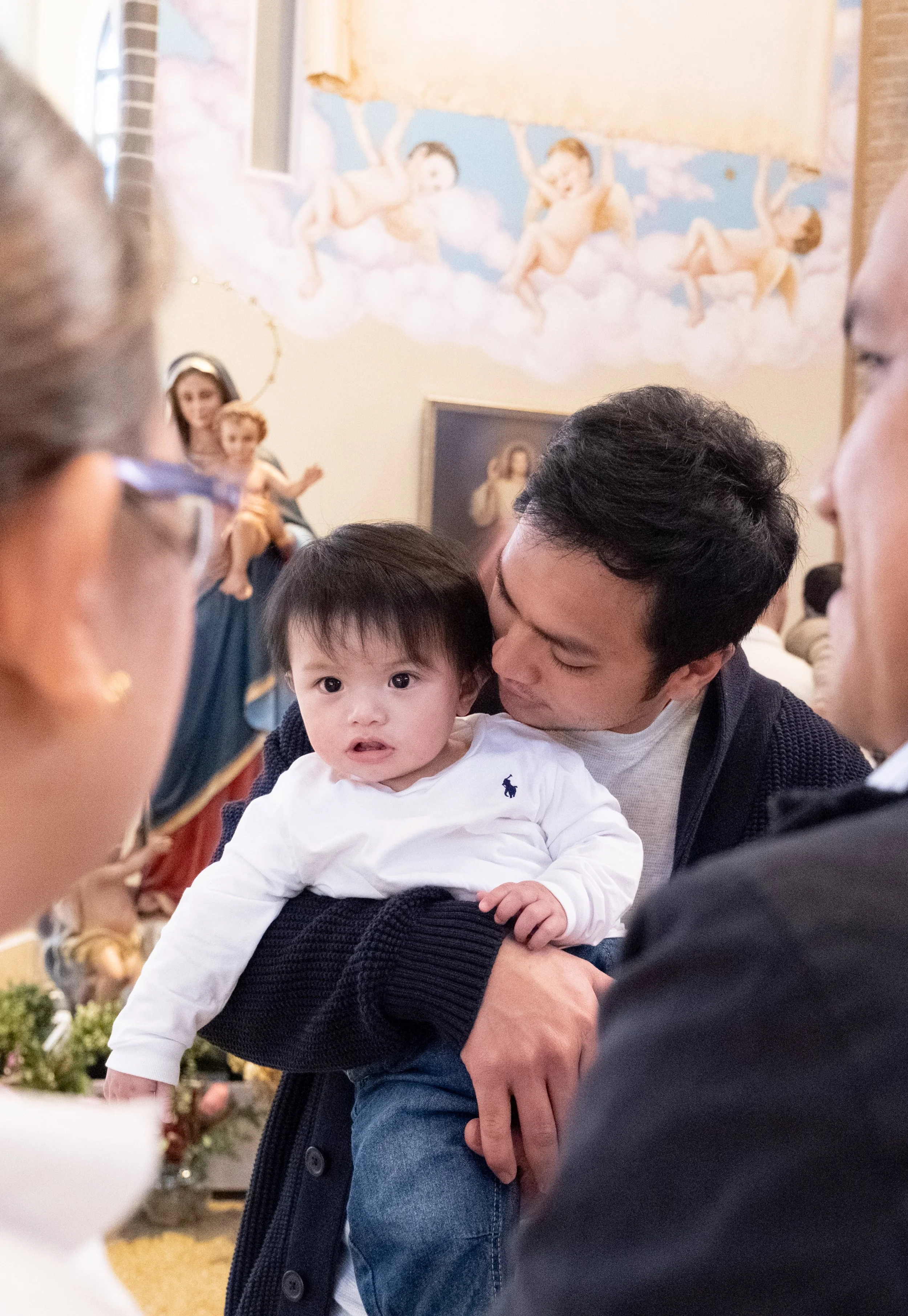 A man holding a young boy during a religious ceremony in a church with religious paintings and a mural of cherubs in the background.