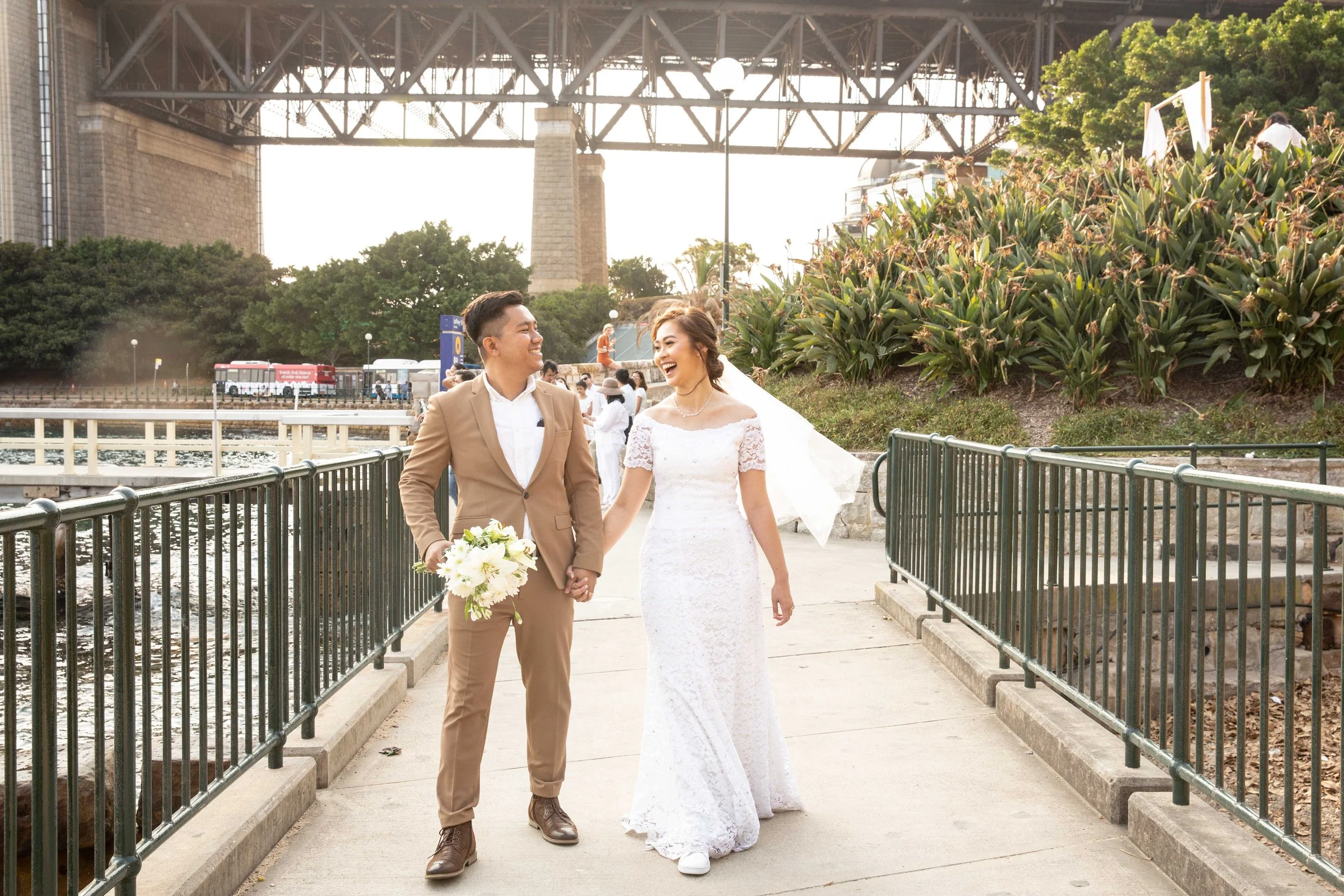 A newly married couple walking hand in hand outdoors near a bridge, smiling and happy. The bride is wearing a white lace wedding dress with a veil, and the groom is dressed in a tan suit with a white shirt. The background features greenery, a waterfr