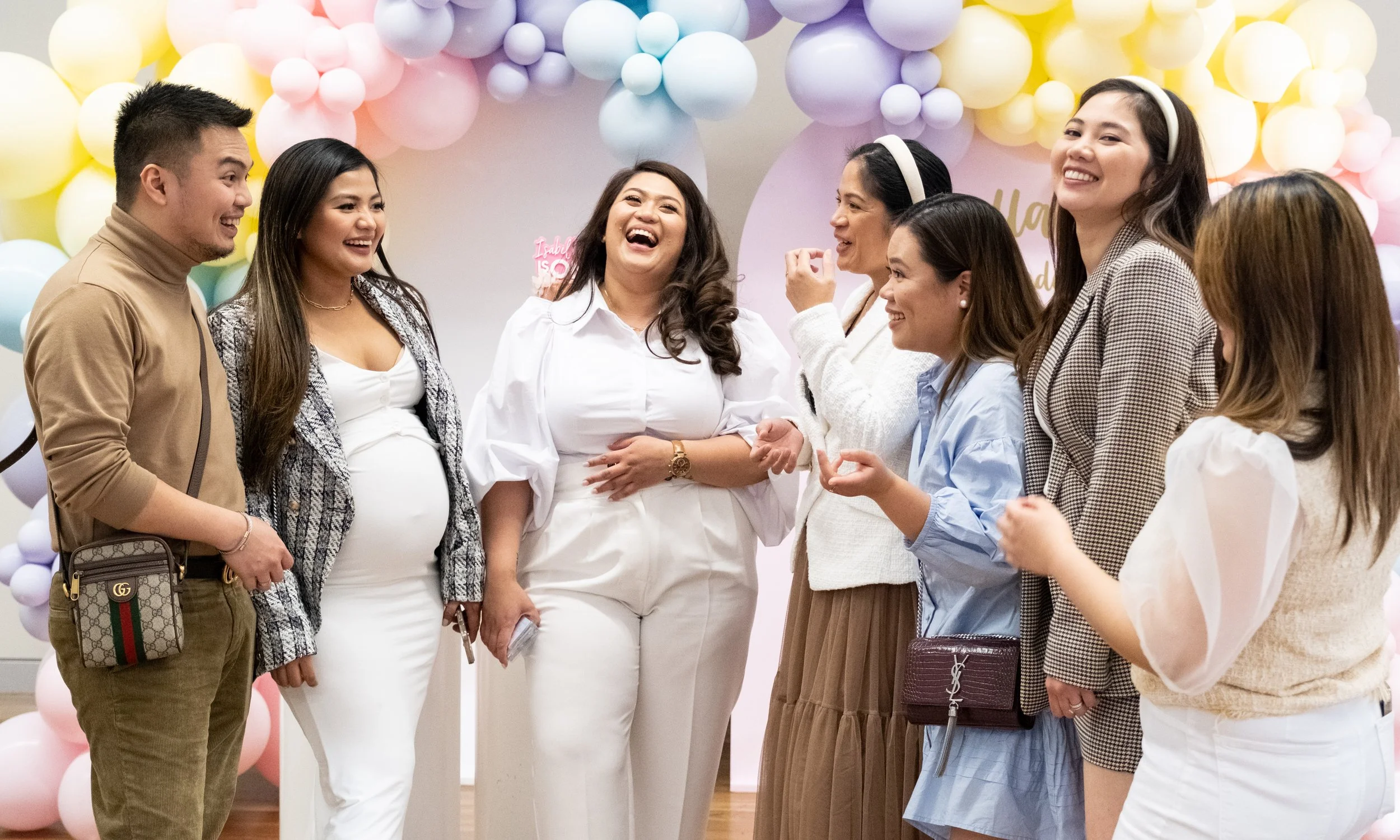 A group of women and a man stand together at a celebration, laughing and talking, with pastel-colored balloon decorations in the background.