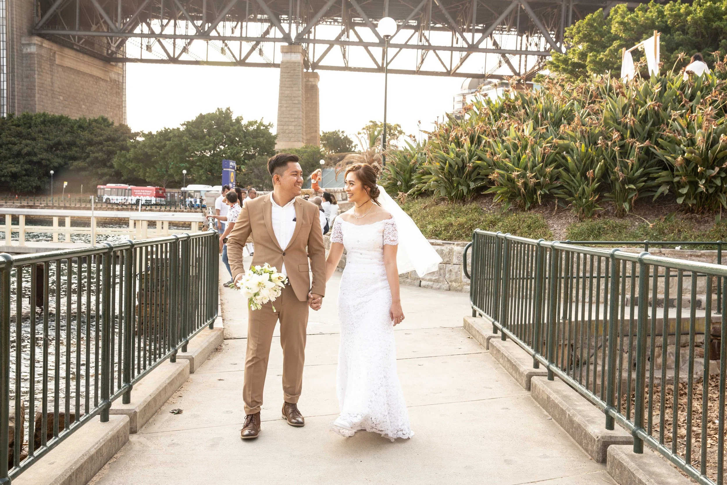 A newlywed couple walking hand-in-hand on a lakeside pathway, smiling at each other, with the Brooklyn Bridge in the background, trees, and onlookers.