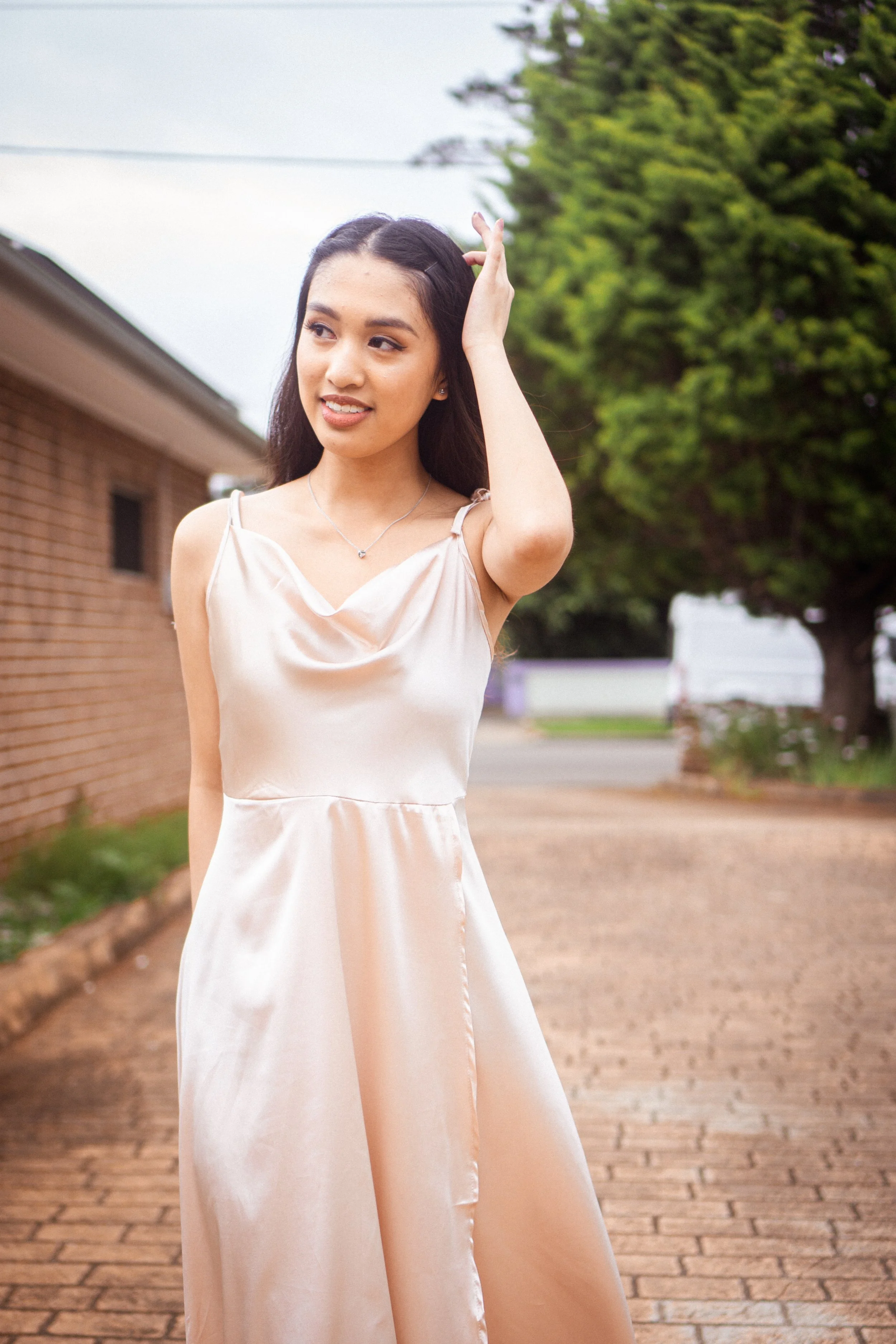 A young woman in a beige satin dress standing outdoors on a brick path, with green trees and a brick building in the background, touching her hair.