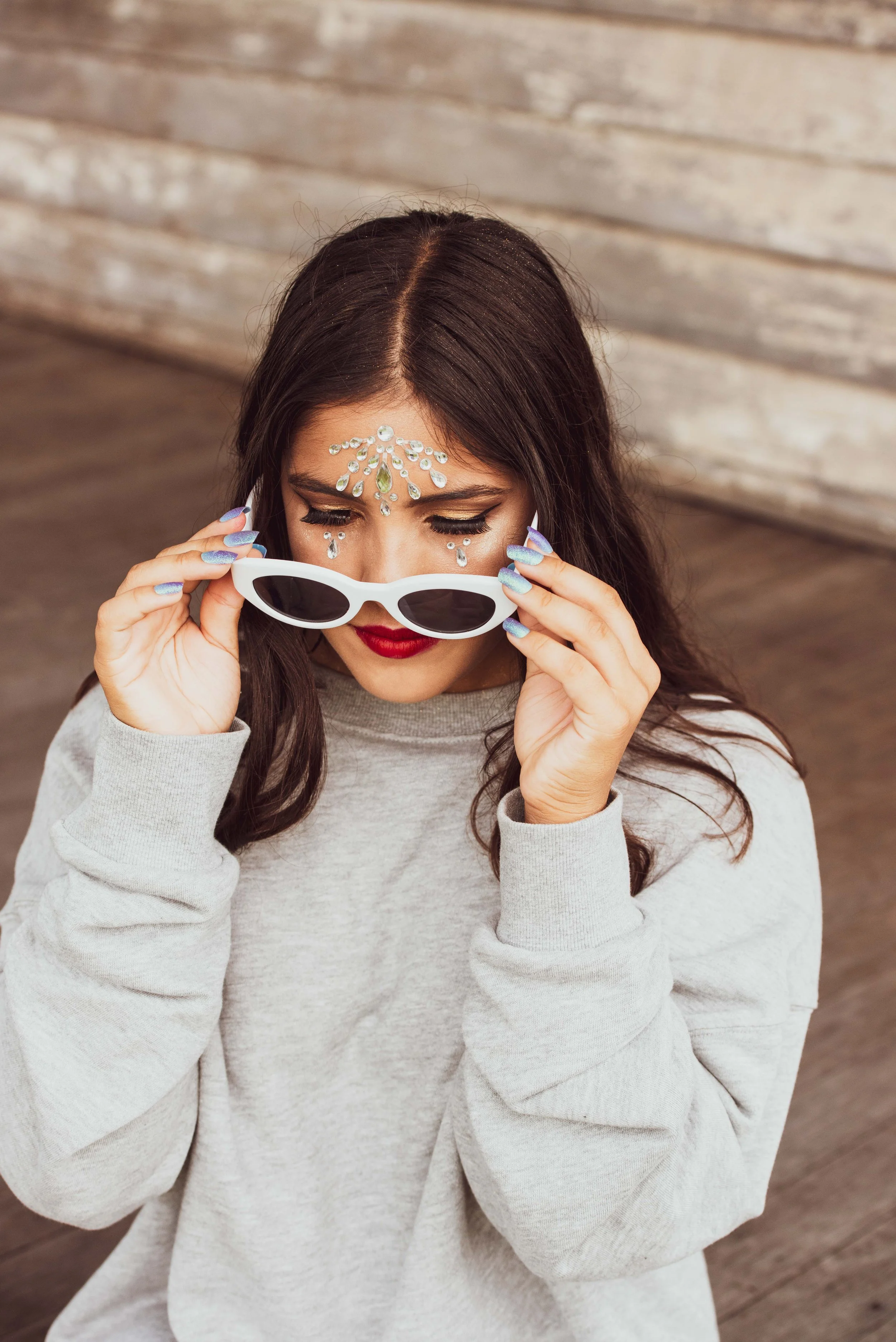 A woman with dark hair and red lipstick holding white sunglasses, wearing decorative rhinestones on her forehead and around her eyes, against a textured wooden background.