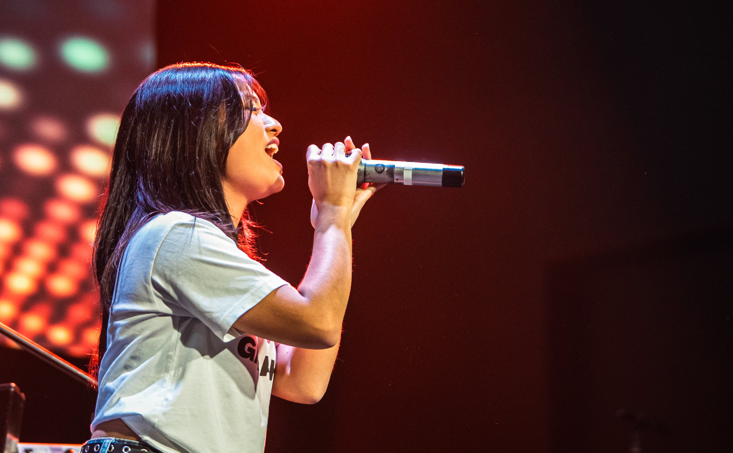 A woman with medium-length dark hair singing into a microphone on stage with colorful lights in the background.