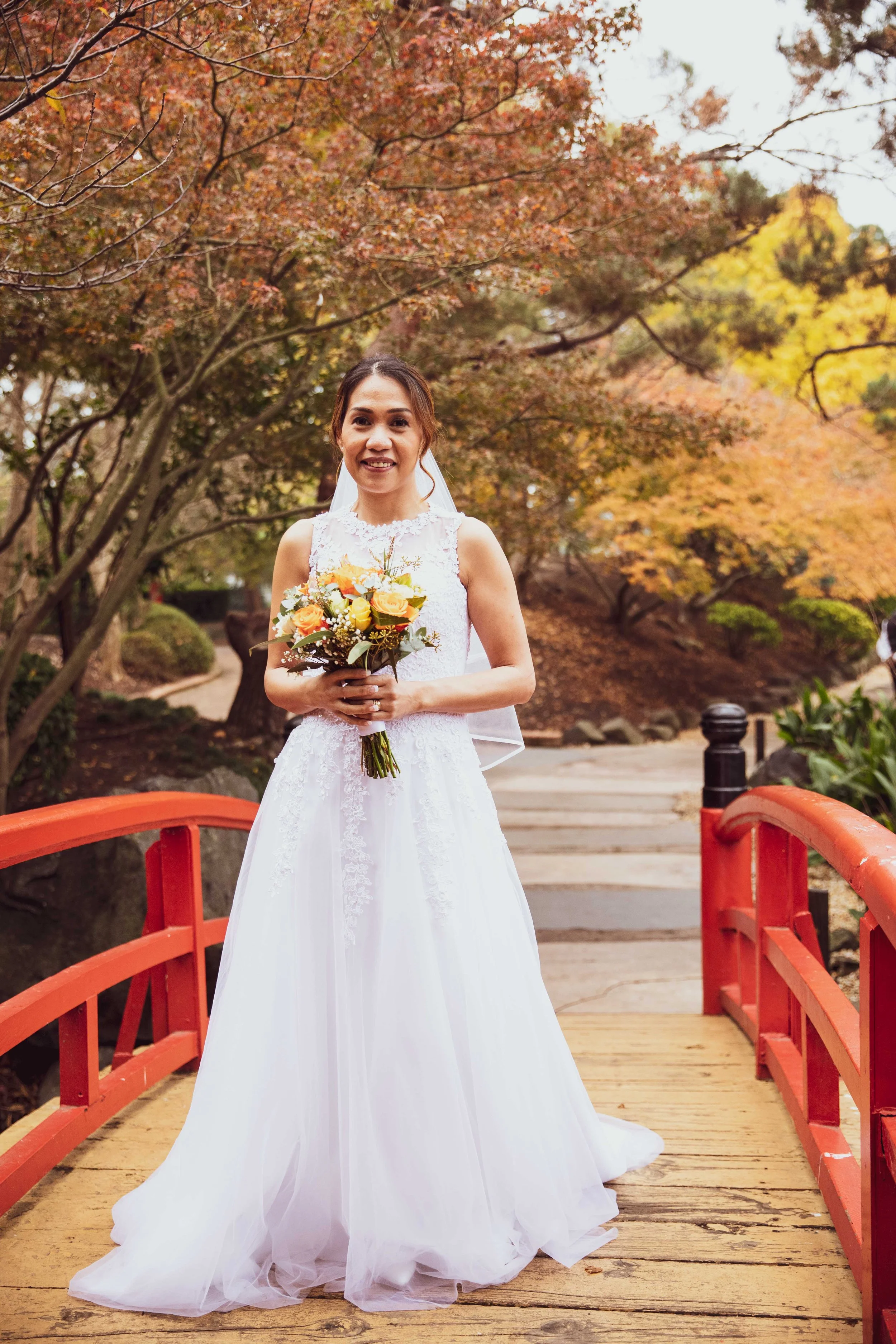 A bride in a white wedding gown holding a bouquet of yellow, orange, and white flowers on a small red wooden bridge surrounded by autumn trees.
