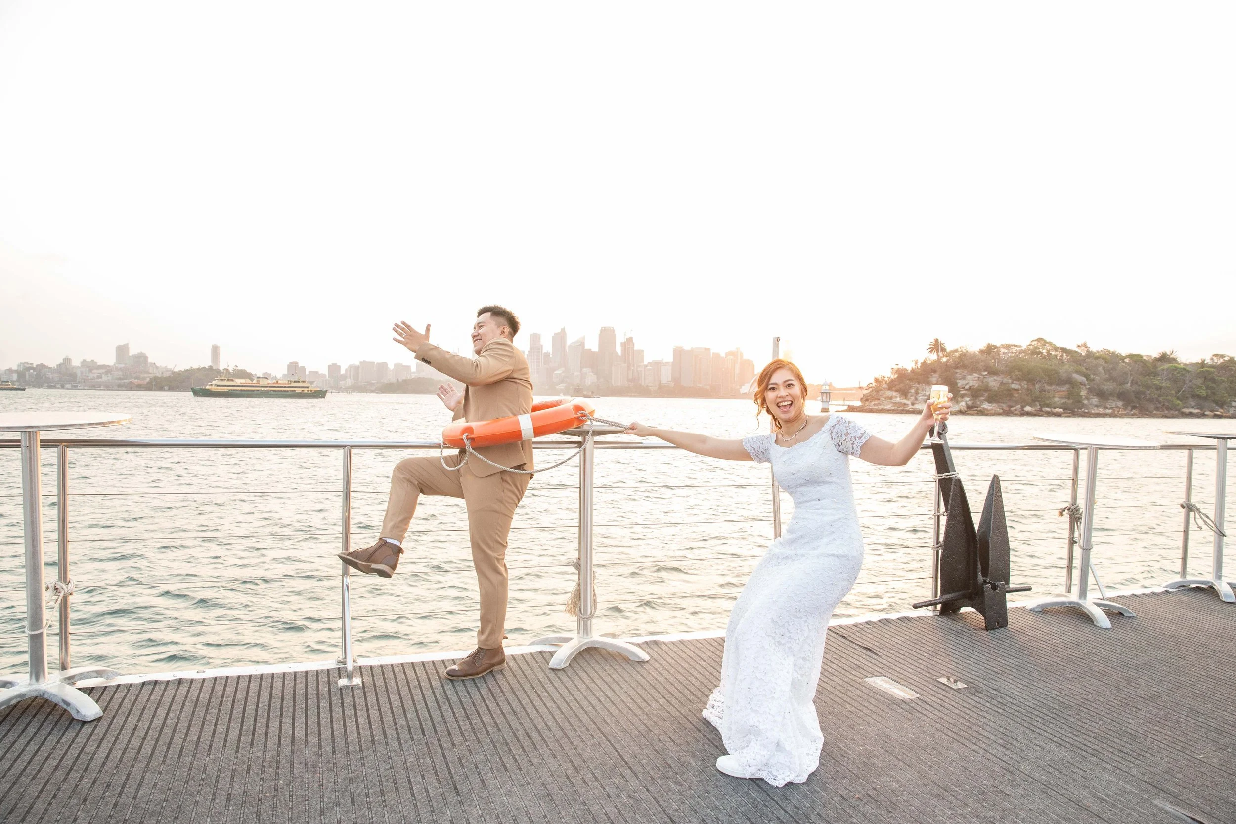 A woman in a white dress and a man in a beige suit on a boat deck with city skyline behind them. The woman is holding a glass of champagne, smiling and pulling the man by his coat, who is pretending to fall into the water while holding onto a life pr