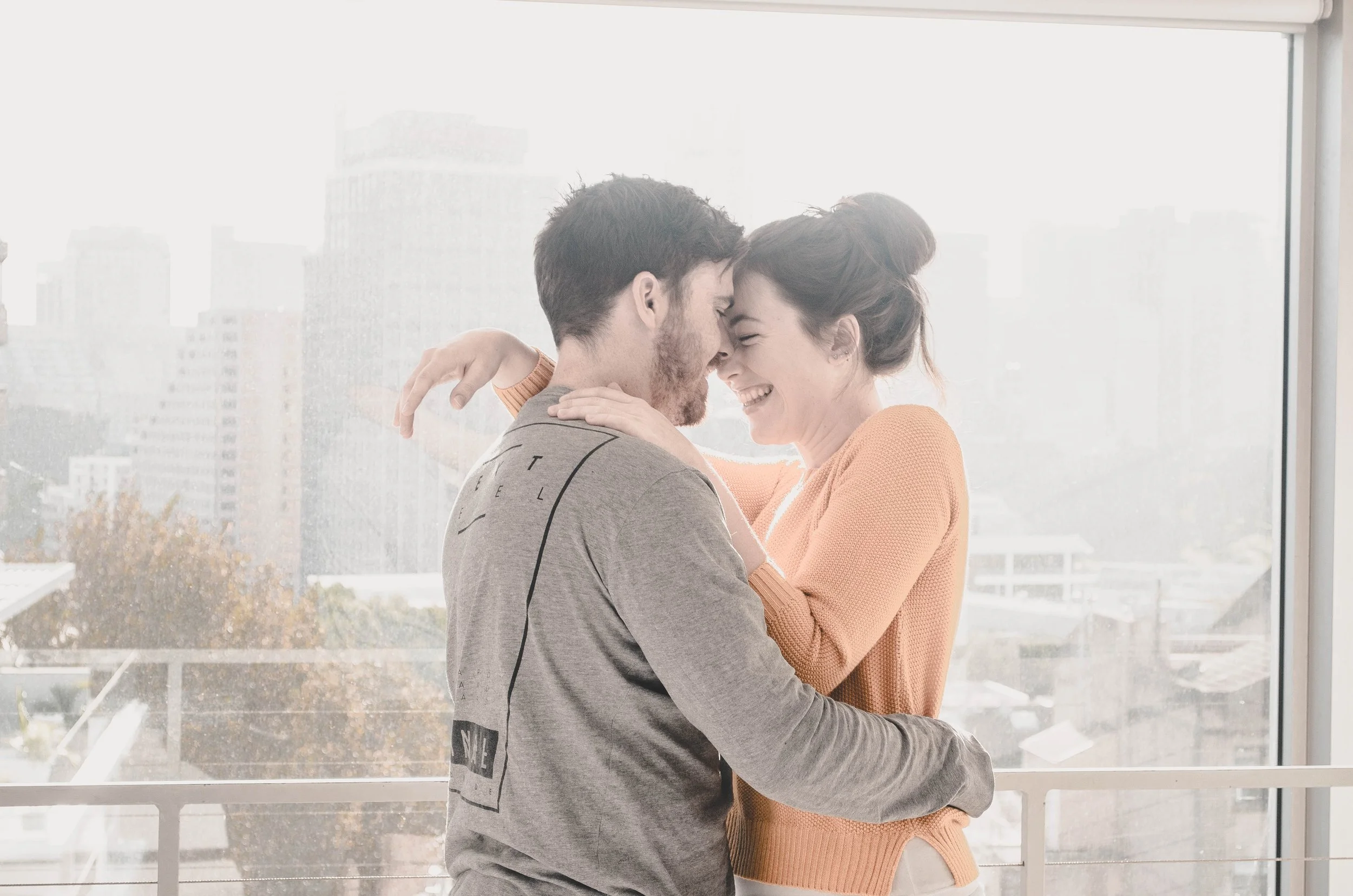 A smiling couple embracing each other, nose to nose, indoors with large windows and city buildings in the background.
