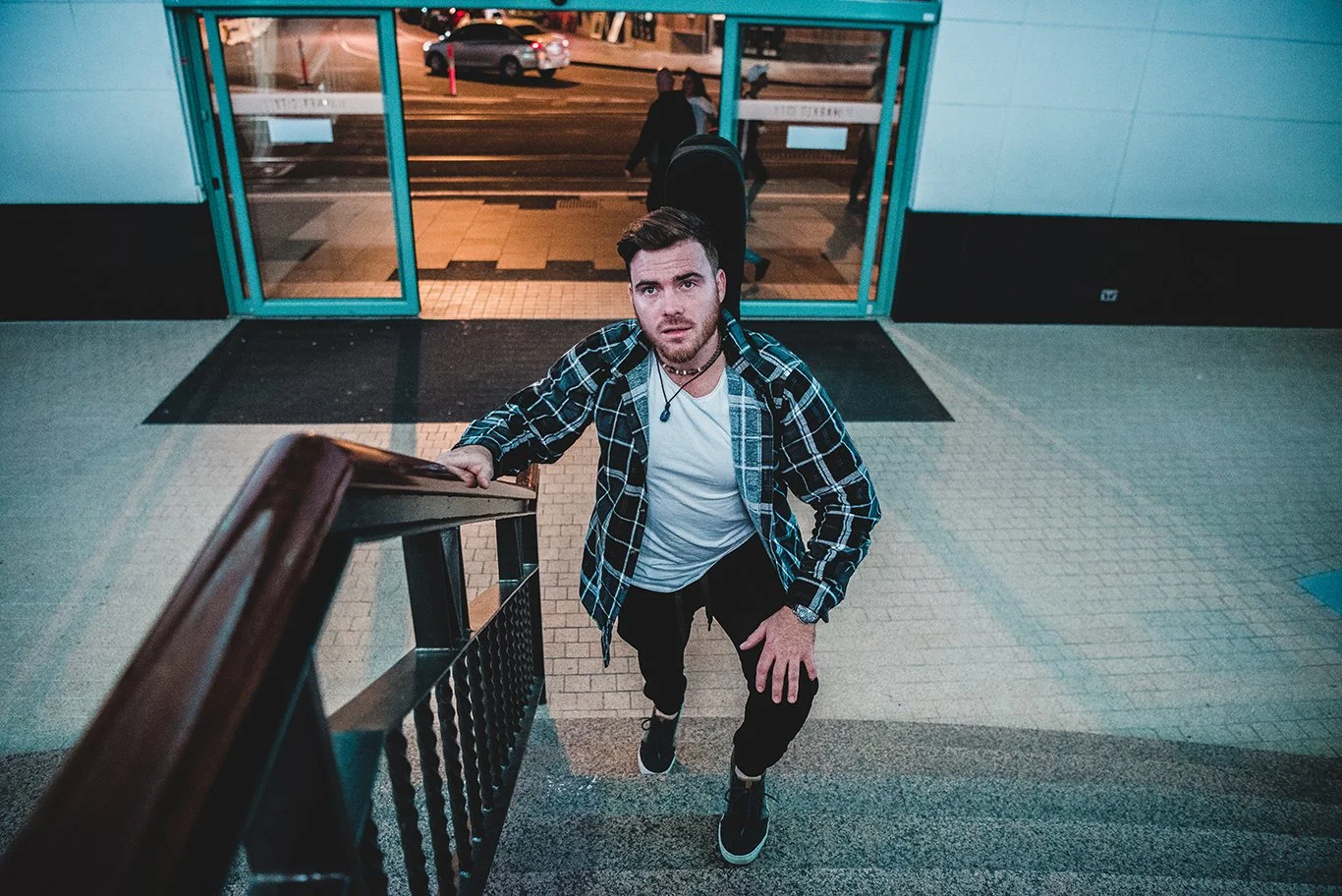 A young man with brown hair, beard, and wearing a plaid shirt, white t-shirt, and black pants, is walking up stairs into a building at night, looking up at the camera outside the entrance.