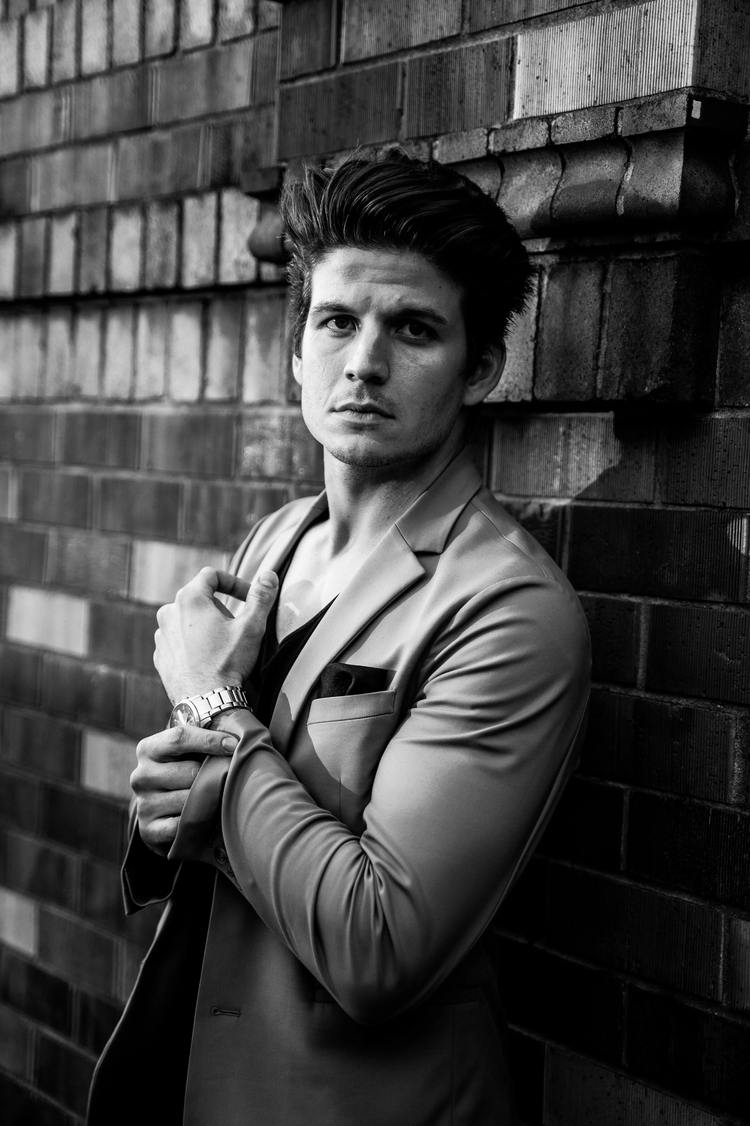 A young man with dark, styled hair, wearing a light-colored blazer and a wristwatch, standing against a brick wall with his arm crossed and looking serious.